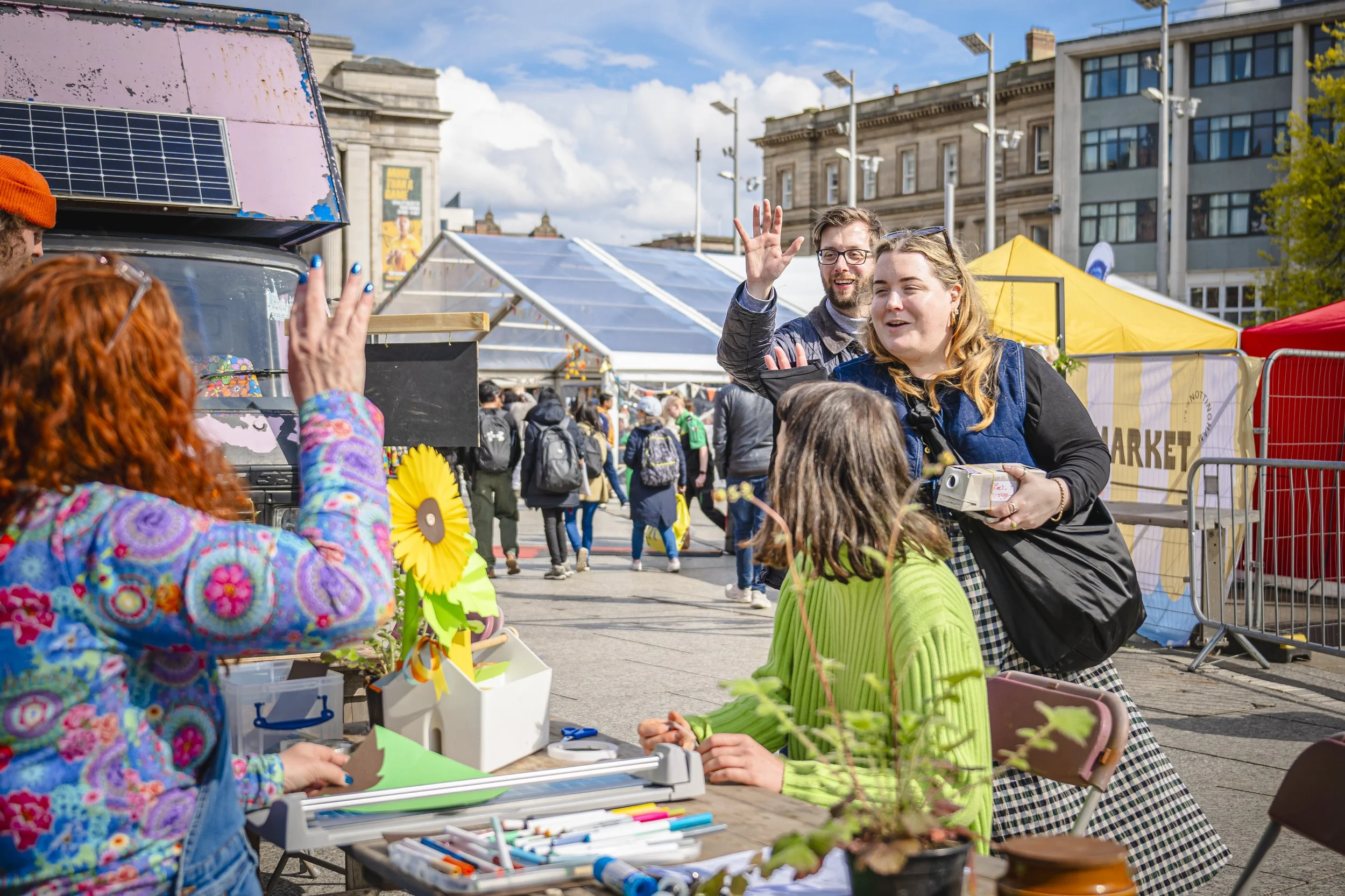 People interacting at an outdoor market with tents and buildings in the background, during the daytime.
