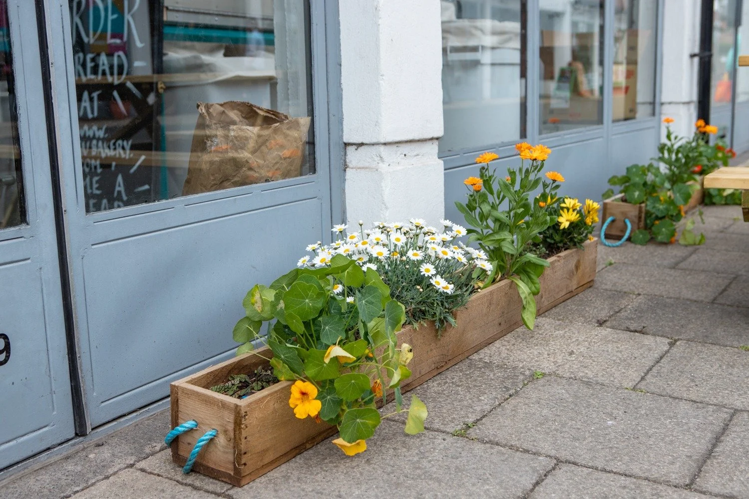 Street view showing wooden planters with colorful flowers outside a storefront with glass windows and light blue frames, on a sidewalk with gray paving stones.