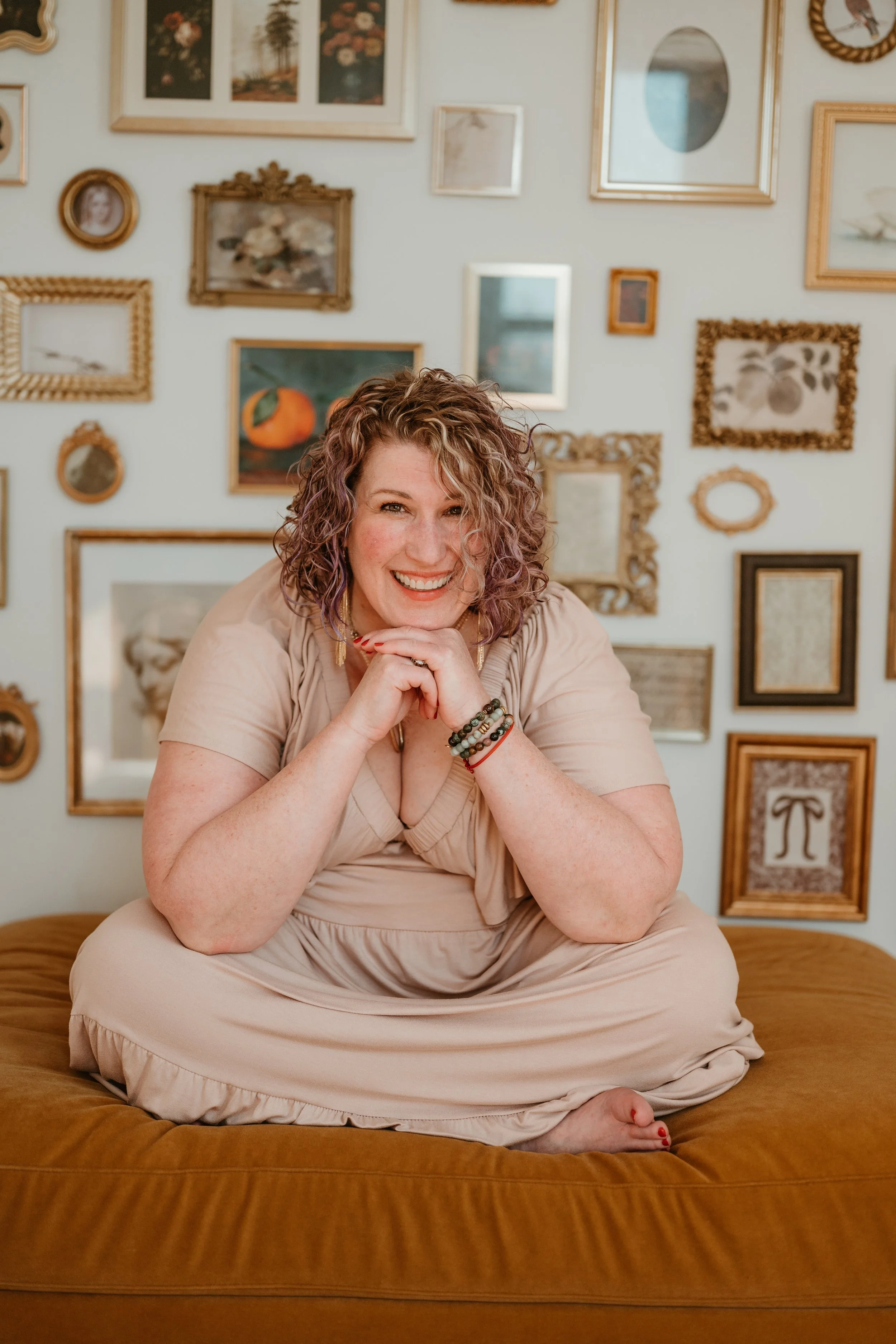 A woman, Page Park, with curly blonde hair, wearing a beige dress, sitting cross-legged on a bed with a brown bench in a room decorated with various framed pictures on the wall behind her. She is smiling and resting her chin on her hands.