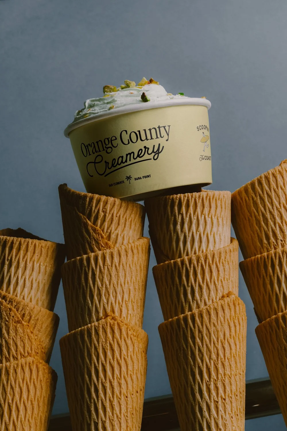 A scoop of vanilla ice cream with pistachio pieces on top, labeled 'Orange County Creamery,' placed on stacked waffle cones.