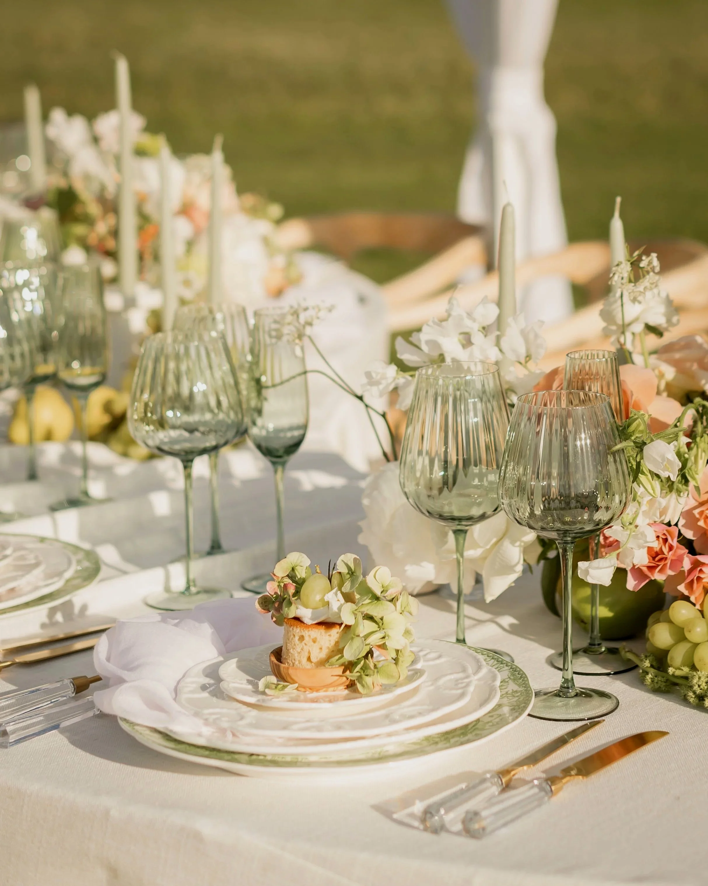 Elegant outdoor table setting with glassware, floral arrangements, candles, and a piece of cake on a plate.