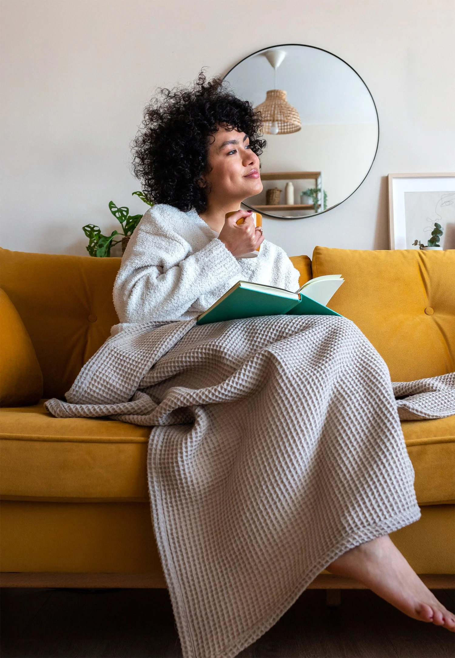 A woman with curly hair sitting on a yellow couch, holding a book and a mug, looking thoughtfully upward in a cozy living room with a mirror, green plants, and framed artwork in the background.