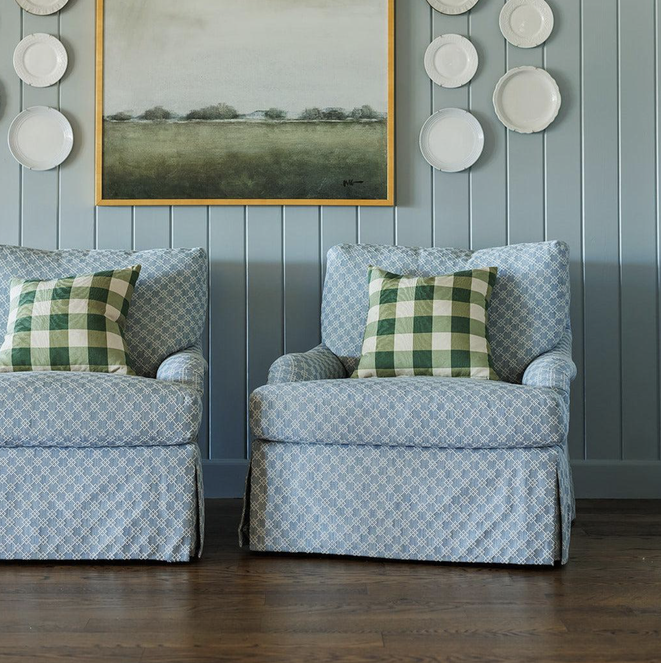 Two blue patterned armchairs with green and white checkered pillows, a painting of a landscape on a blue paneled wall, and decorative white plates on the wall.