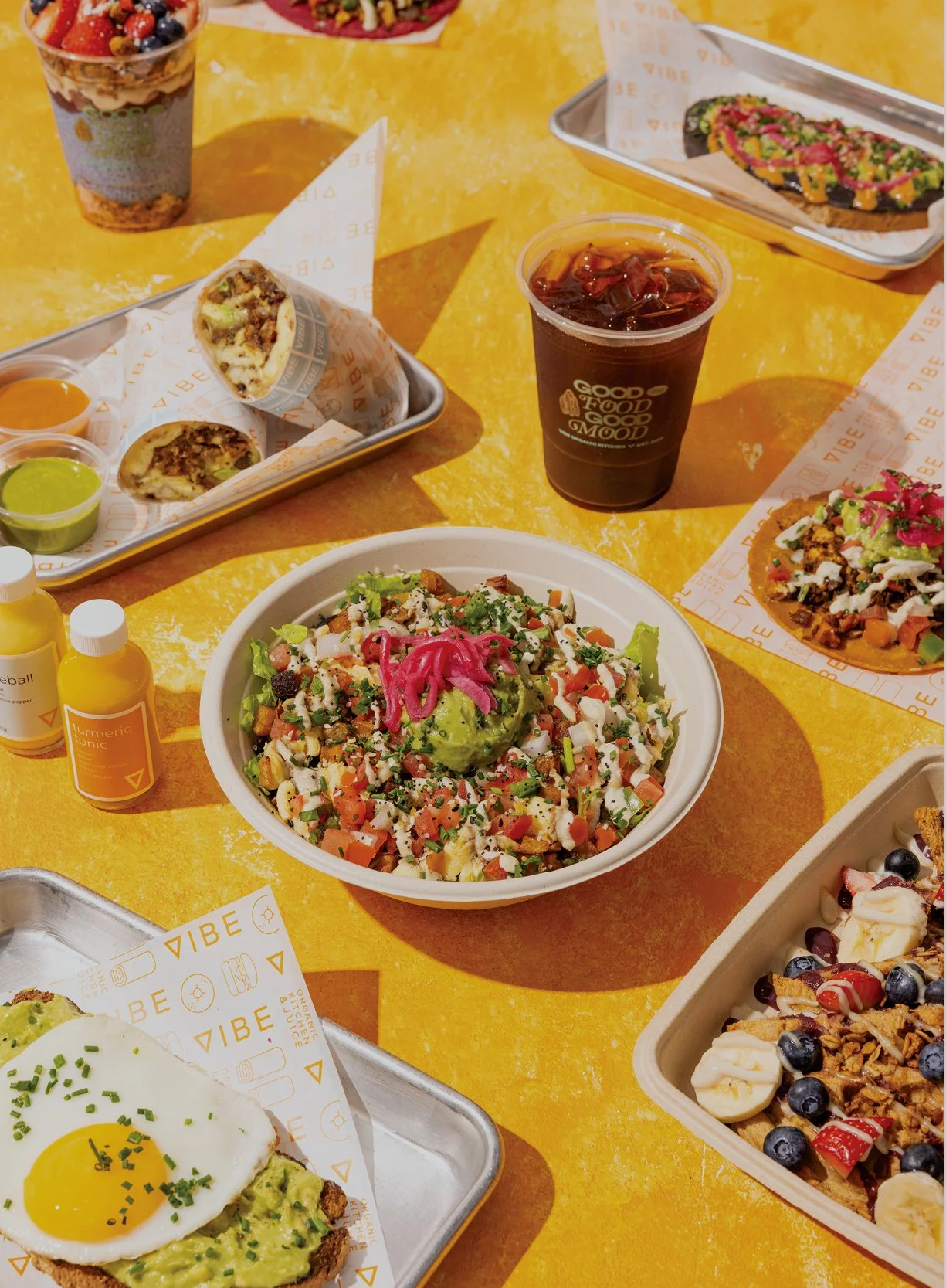 Colorful meal spread on a yellow table including a salad bowl with avocado, vegetables, and pickled pink onions, a pizza with greens and vegetables, a desert with berries and bananas, a cup of iced coffee, small bottles of turmeric tonic, and wraps and tacos with various ingredients.