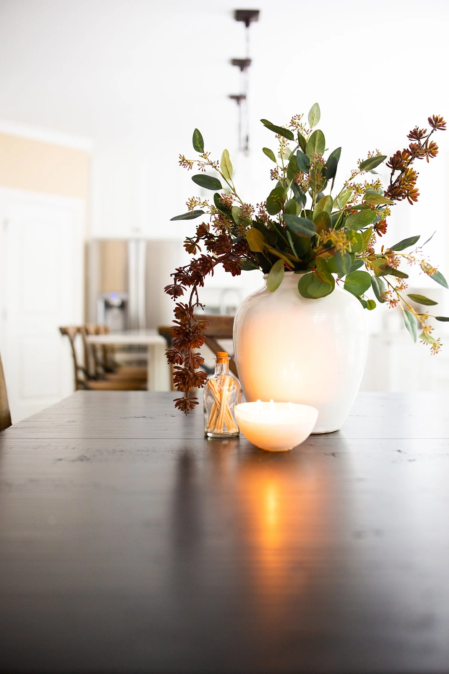 A white vase with green and brown foliage, a lit candle, and a small bottle with wooden sticks on a dark wooden dining table in a bright room.
