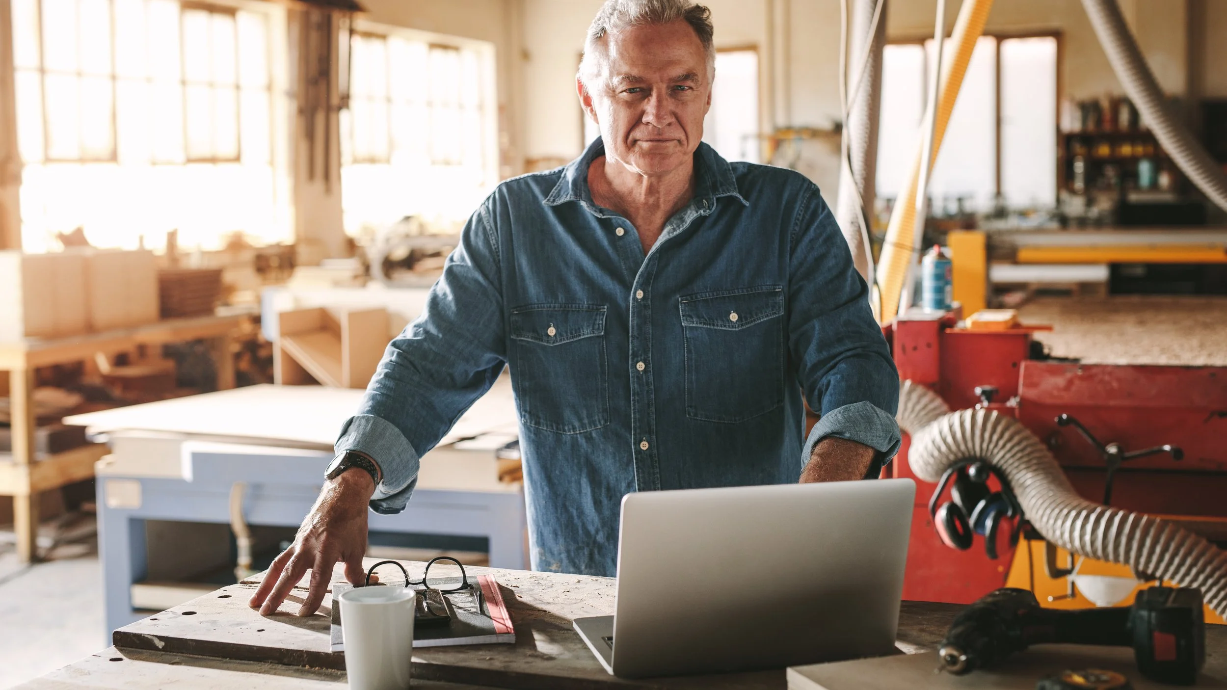 A man wearing a denim shirt working in a woodworking shop with a laptop, glasses, and a cup on the table.