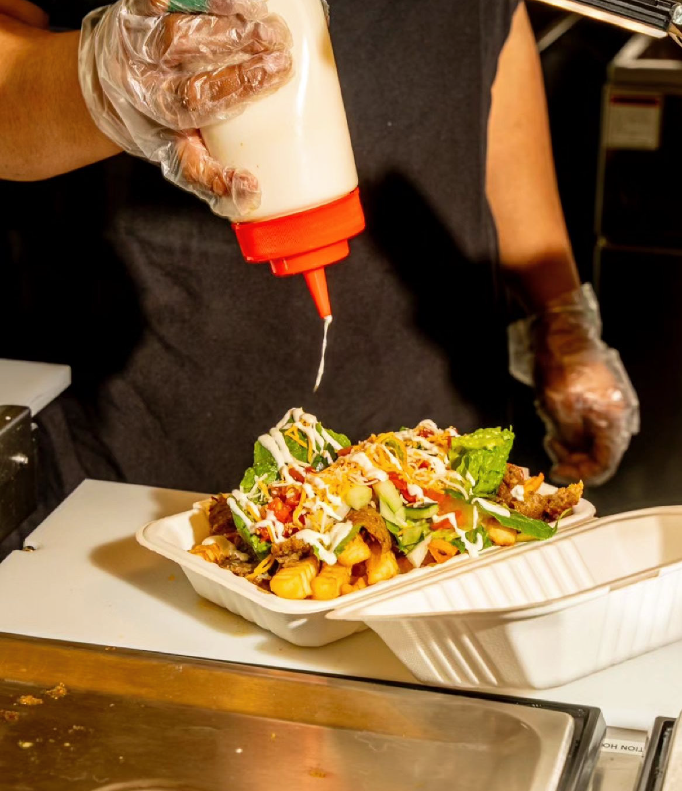 Person wearing gloves pouring mayonnaise onto a serving of loaded fries topped with lettuce, shredded cheese, and vegetables.