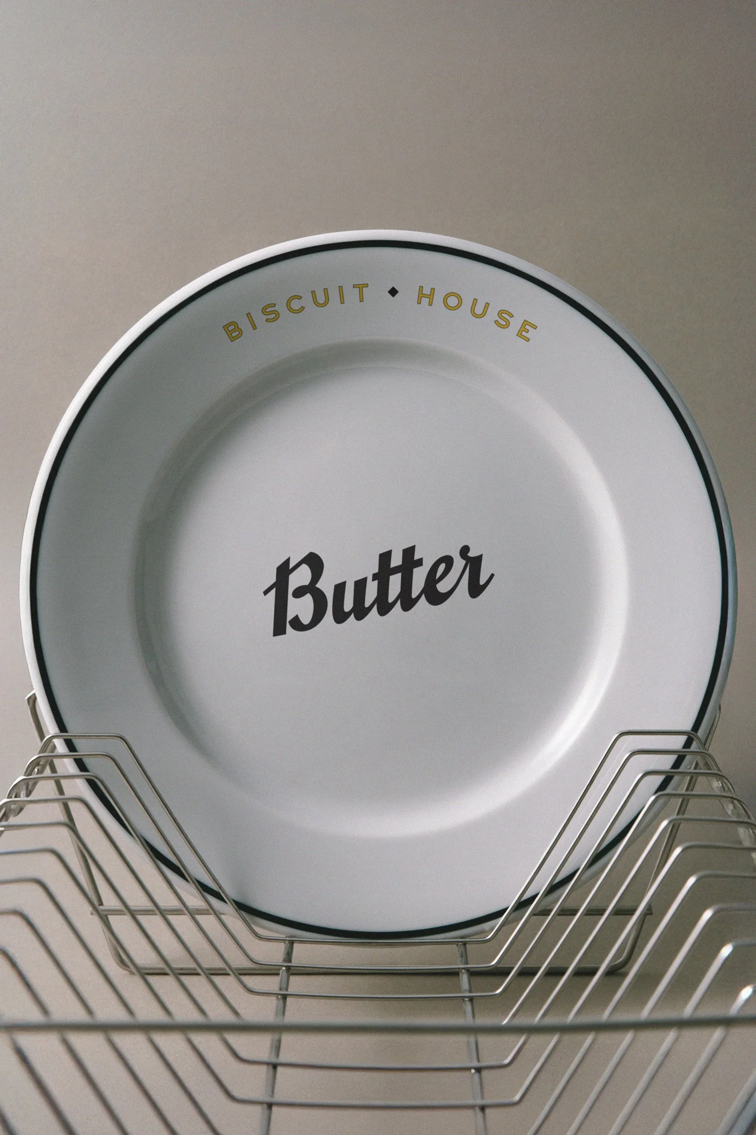 White ceramic plate with the word 'Butter' in black script, rimmed with black and gold text reading 'Biscuit House', placed on a wire dish rack against a plain light-colored background.