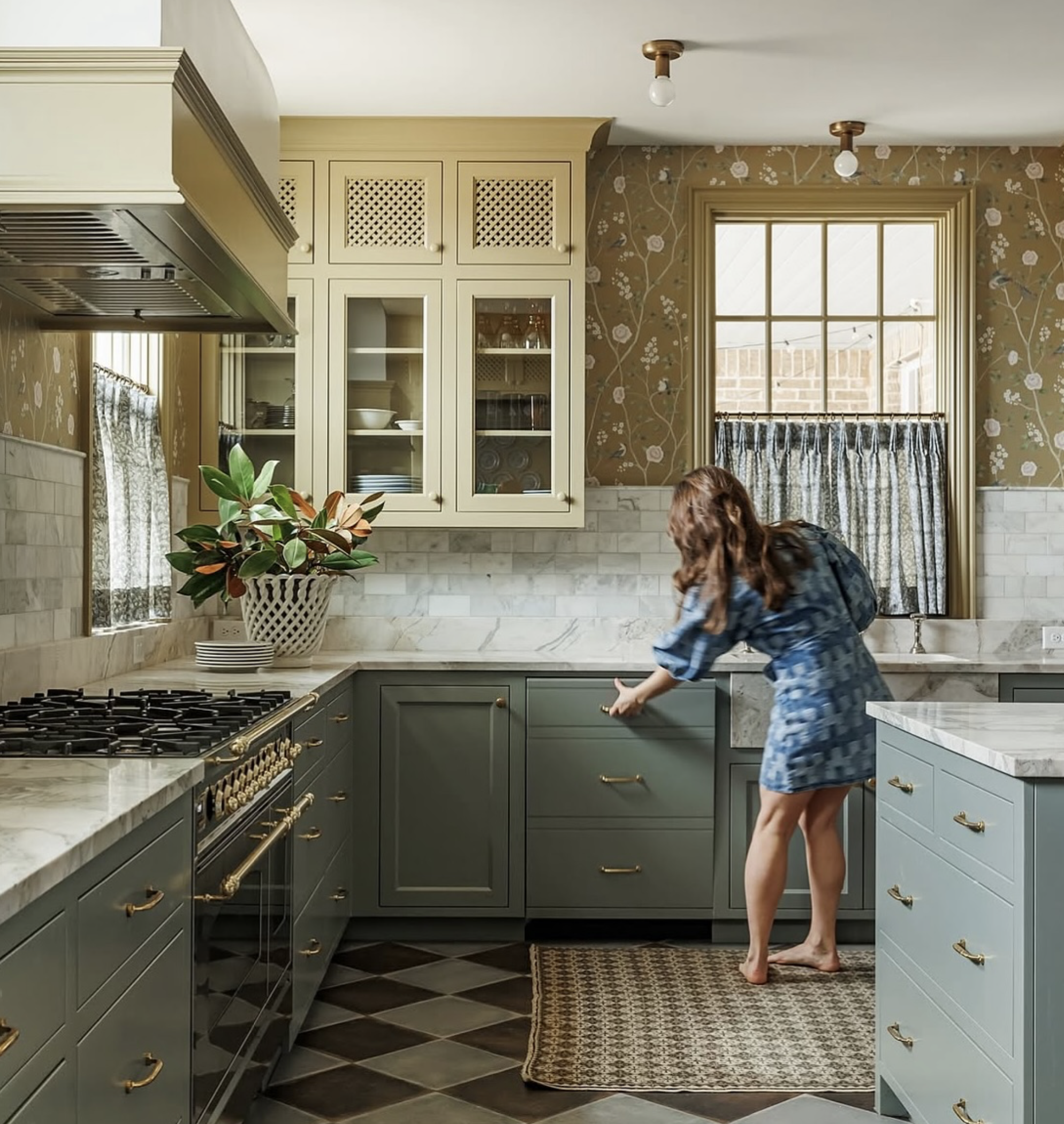 A woman in a blue dress reaching for a drawer in a kitchen with green cabinets, marble countertops, and floral wallpaper, near a window.