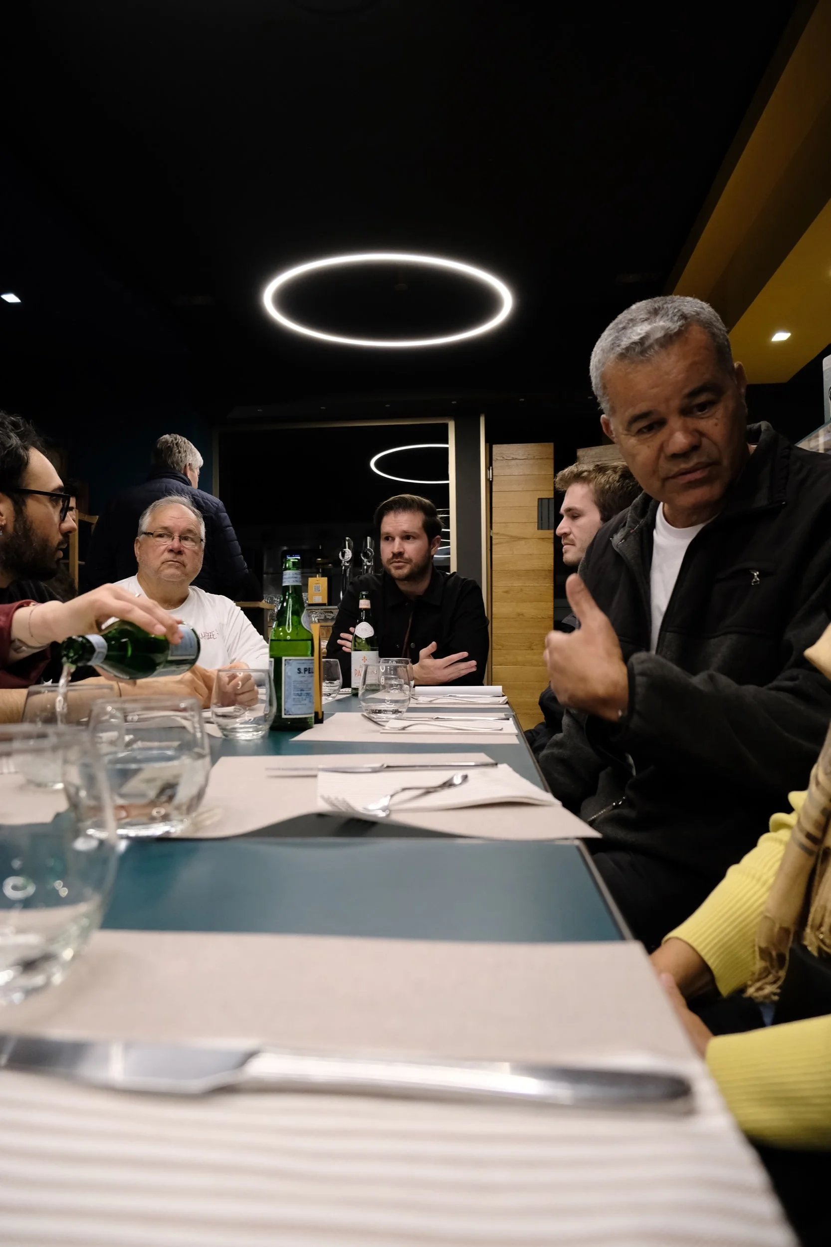 A group of people sitting at a dining table in a restaurant, engaged in conversation. The table has glasses and bottles, and the room has modern circular ceiling lights and a wooden wall.