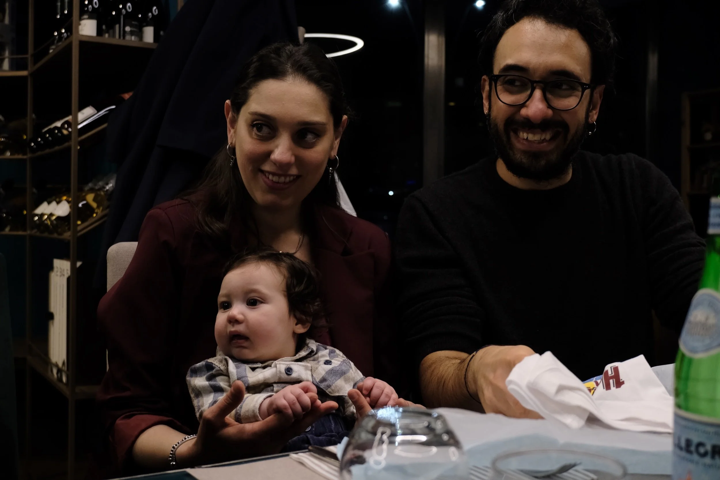 A group of people, including a woman, a man, and a baby, sitting around a table in a dimly lit room with wine racks in the background. The woman and man are smiling, and the baby is looking forward with curiosity.