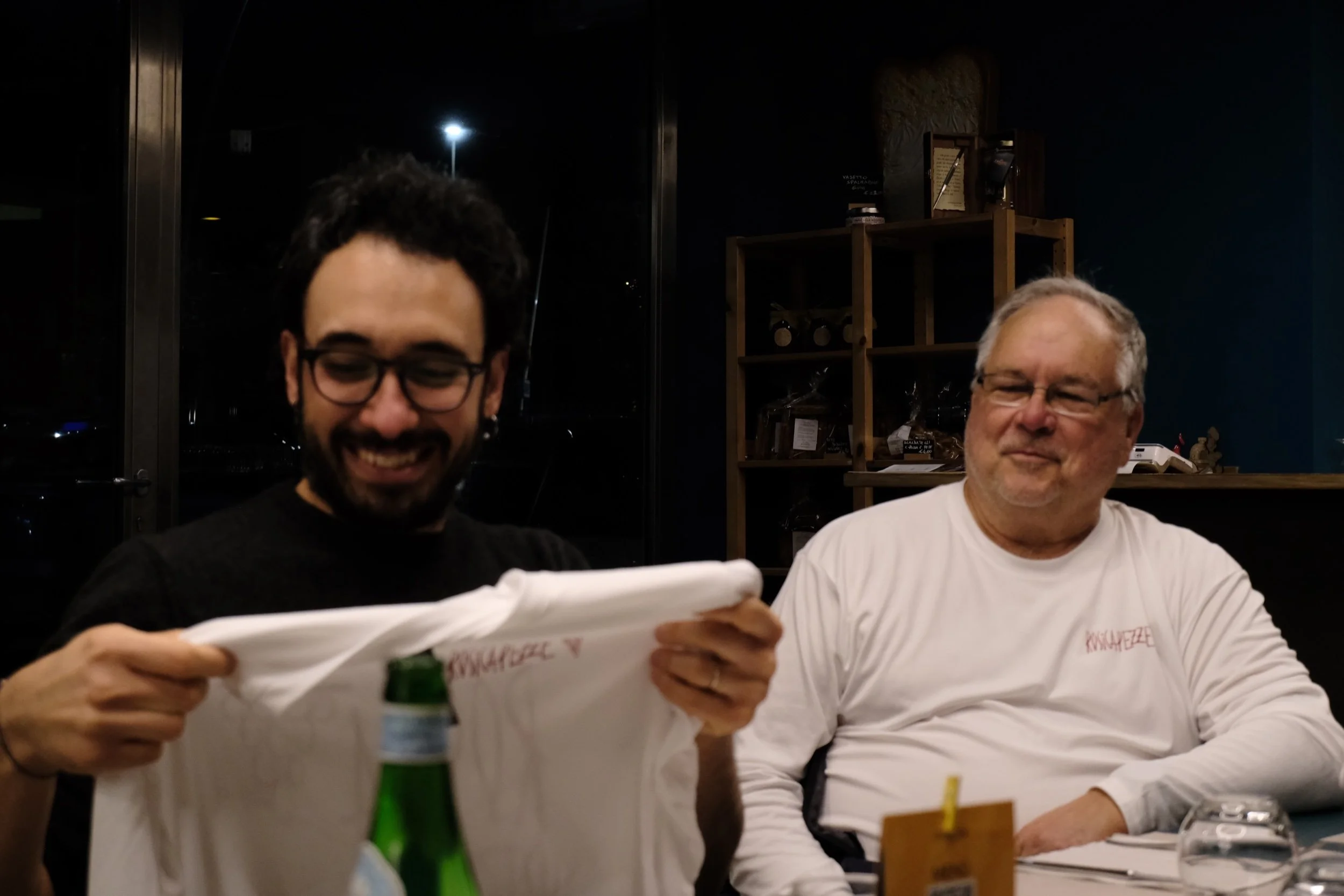 Two men sitting at a table with one holding a clipboard, in a dimly lit room with shelves in the background, possibly in a restaurant or cafe setting.