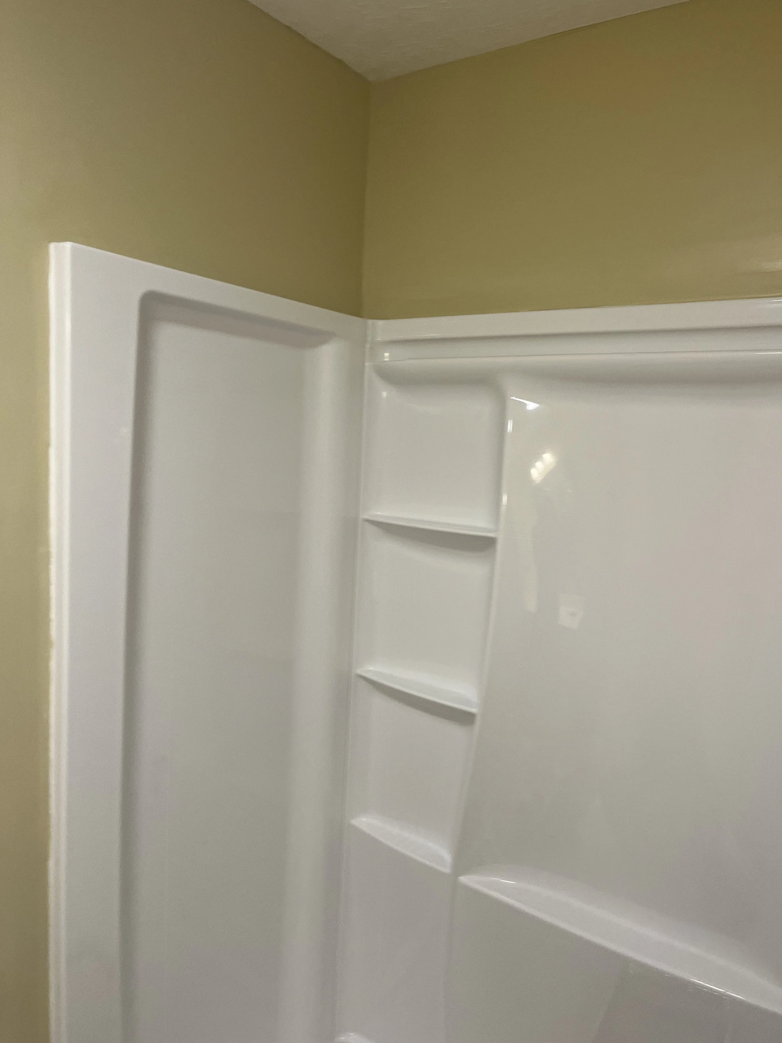 Close-up of a corner of a white shower with shelves and a beige wall in a bathroom.