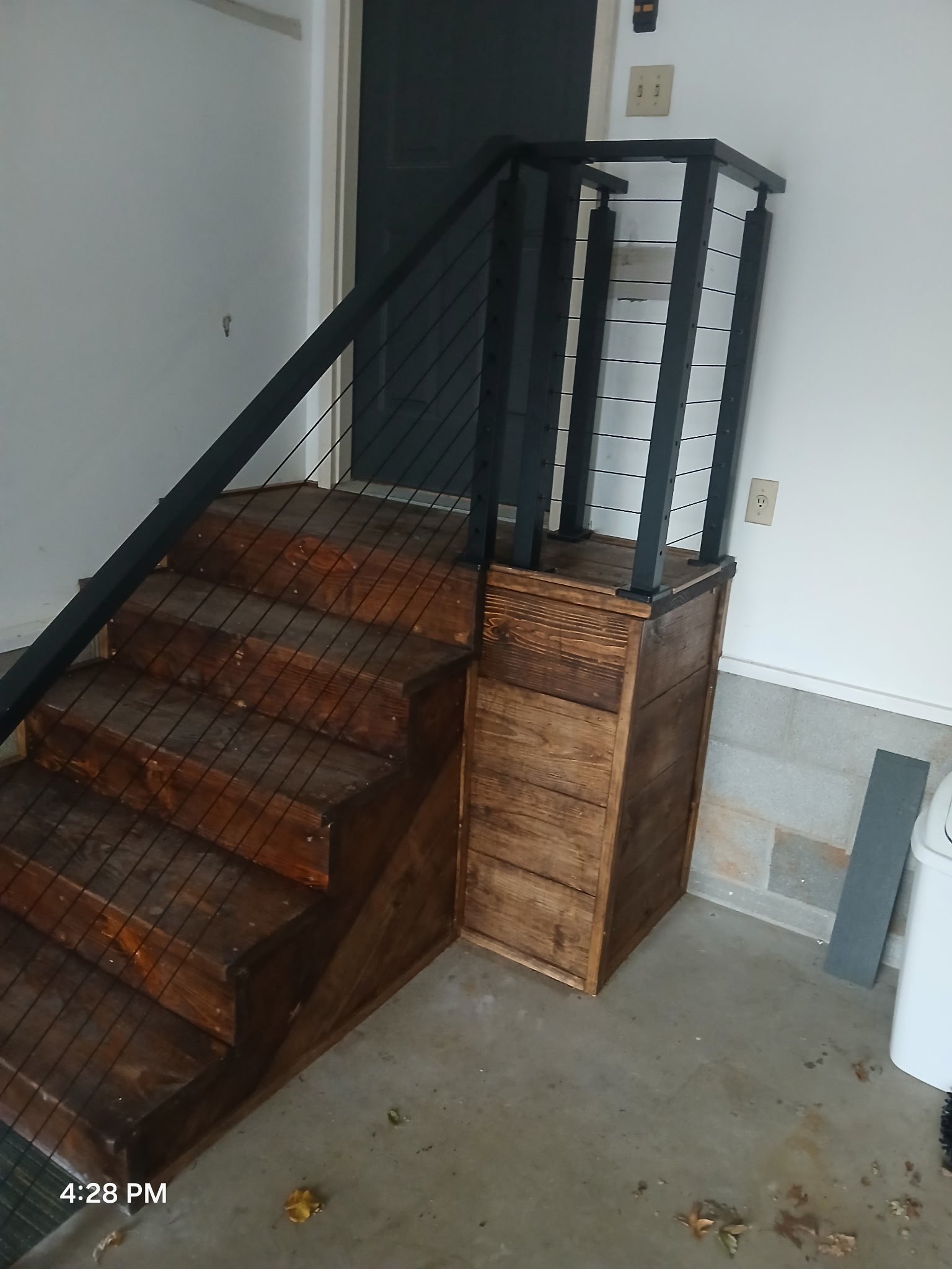 Wooden staircase with black metal railing in a garage or basement with concrete floor, white walls, and a gray door in the background.
