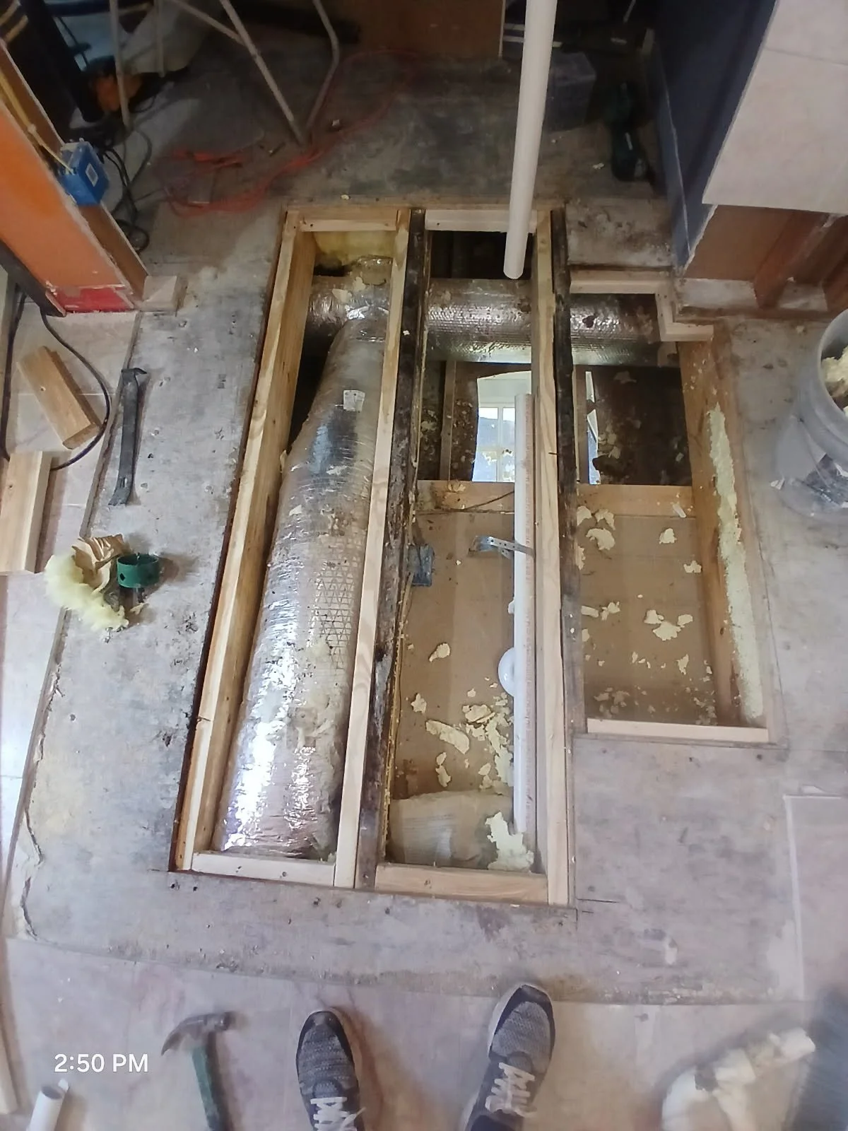 View of a bathroom renovation with exposed wooden framing, plumbing pipes, and insulation, showing the subfloor with some removed tiles.
