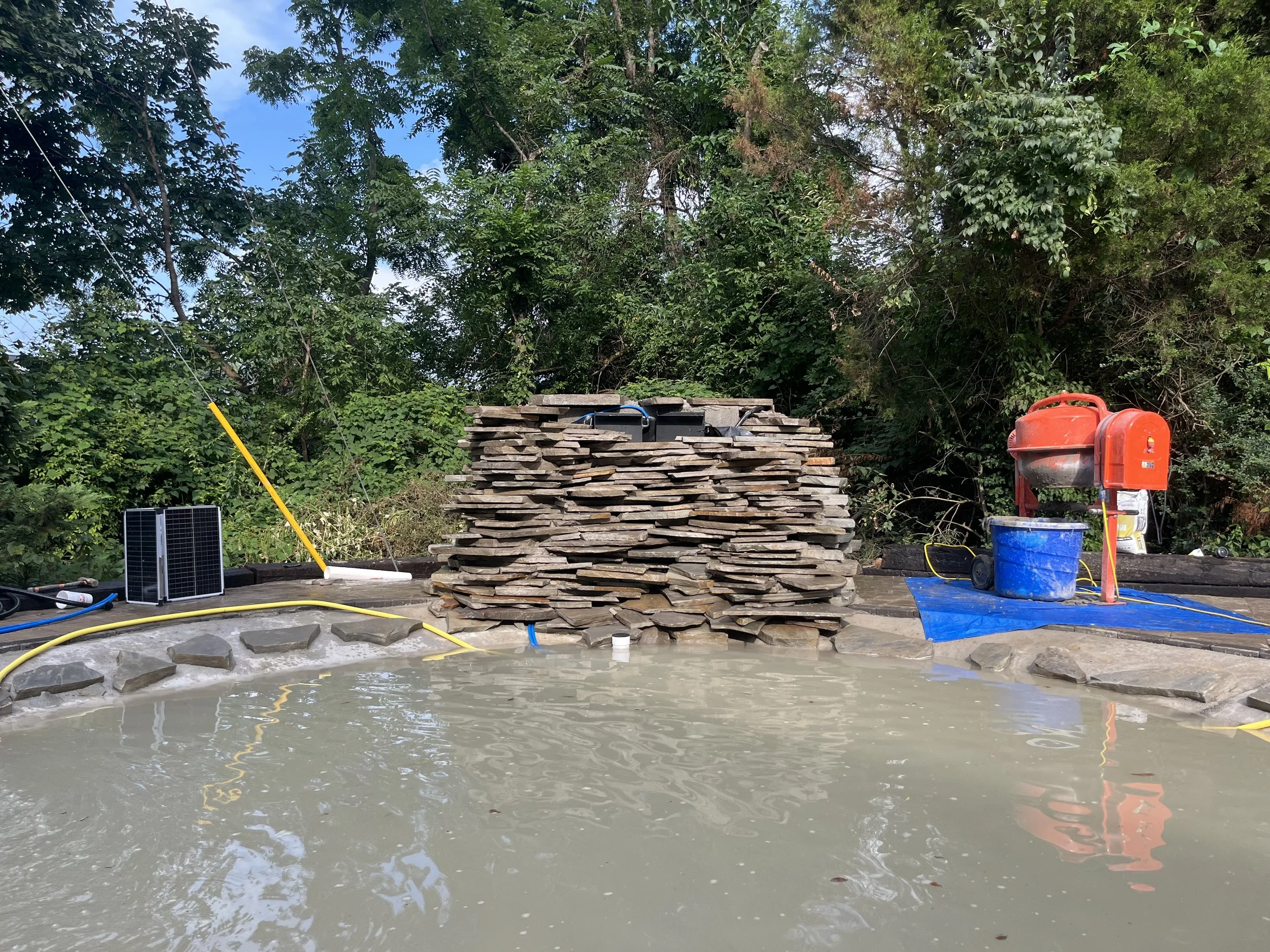 A hot tub under construction with a large pile of flat stones stacked behind it. On the left side, there is a small solar panel and some hoses. On the right, there is a red pump, a blue bucket, and a blue tarp, with a green tree and bushes in the bac