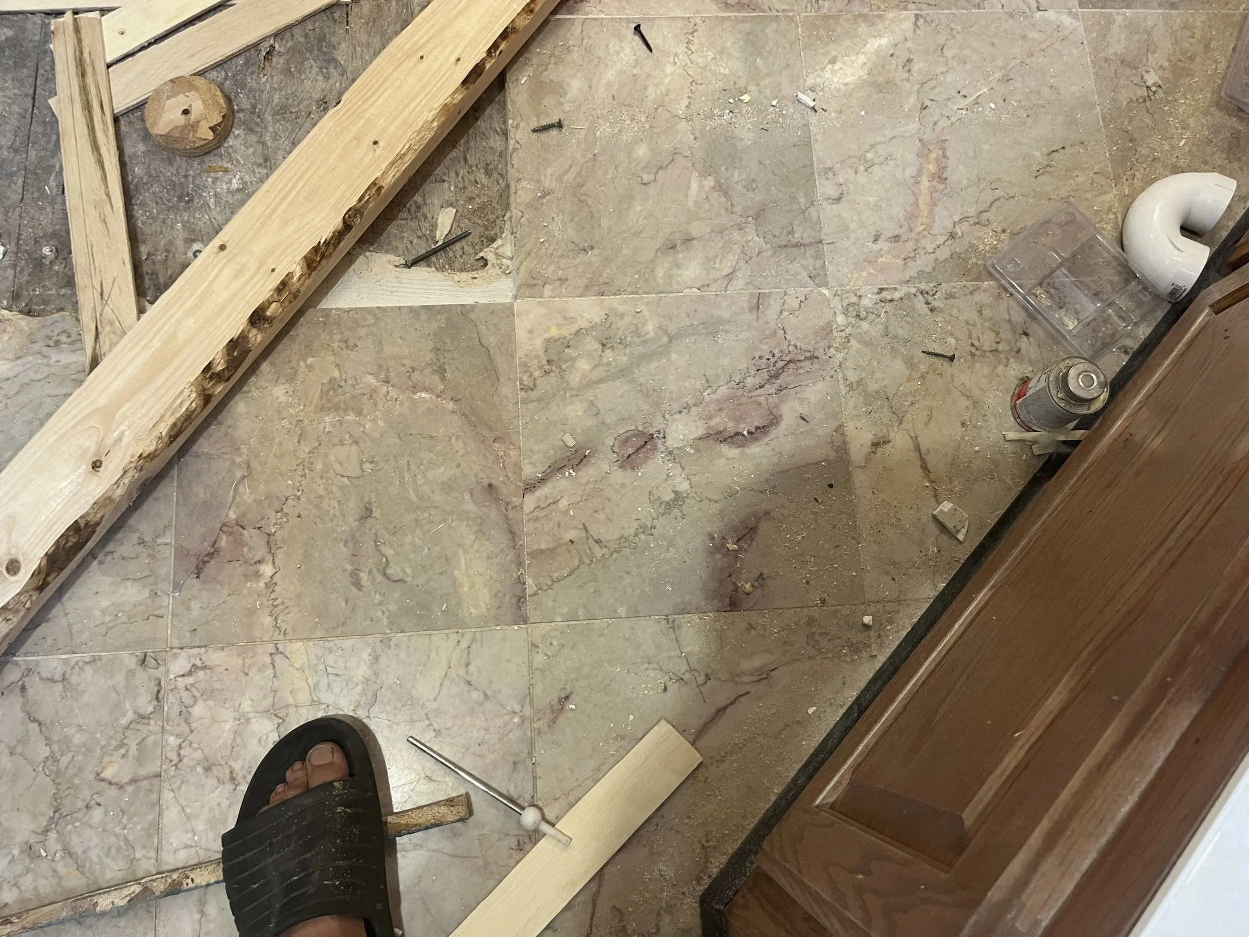 Part of a floor under construction with scattered tools and materials, including a wooden plank, a metal rod, a small level, a white pipe, a spray can, and a clear plastic container. The floor has tiles and exposed subflooring.