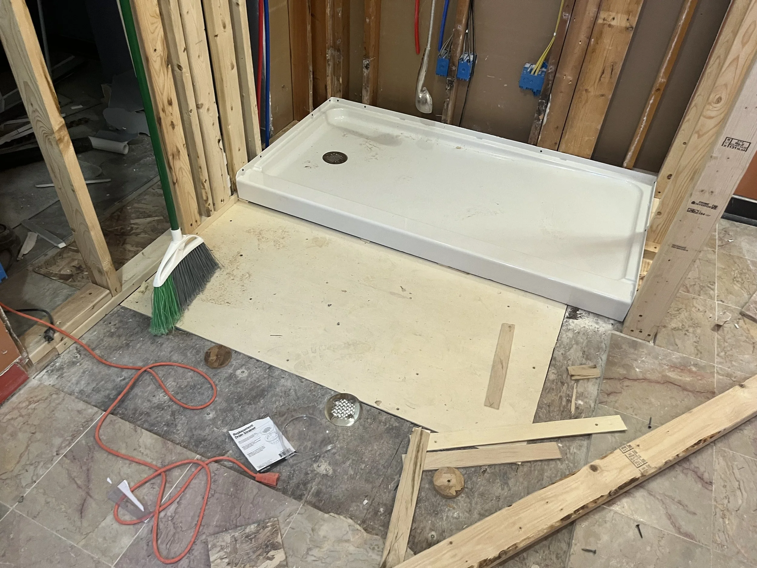 Construction site with a white shower base on a beige sheet, surrounded by wooden framing, a broom, wires, and construction debris.