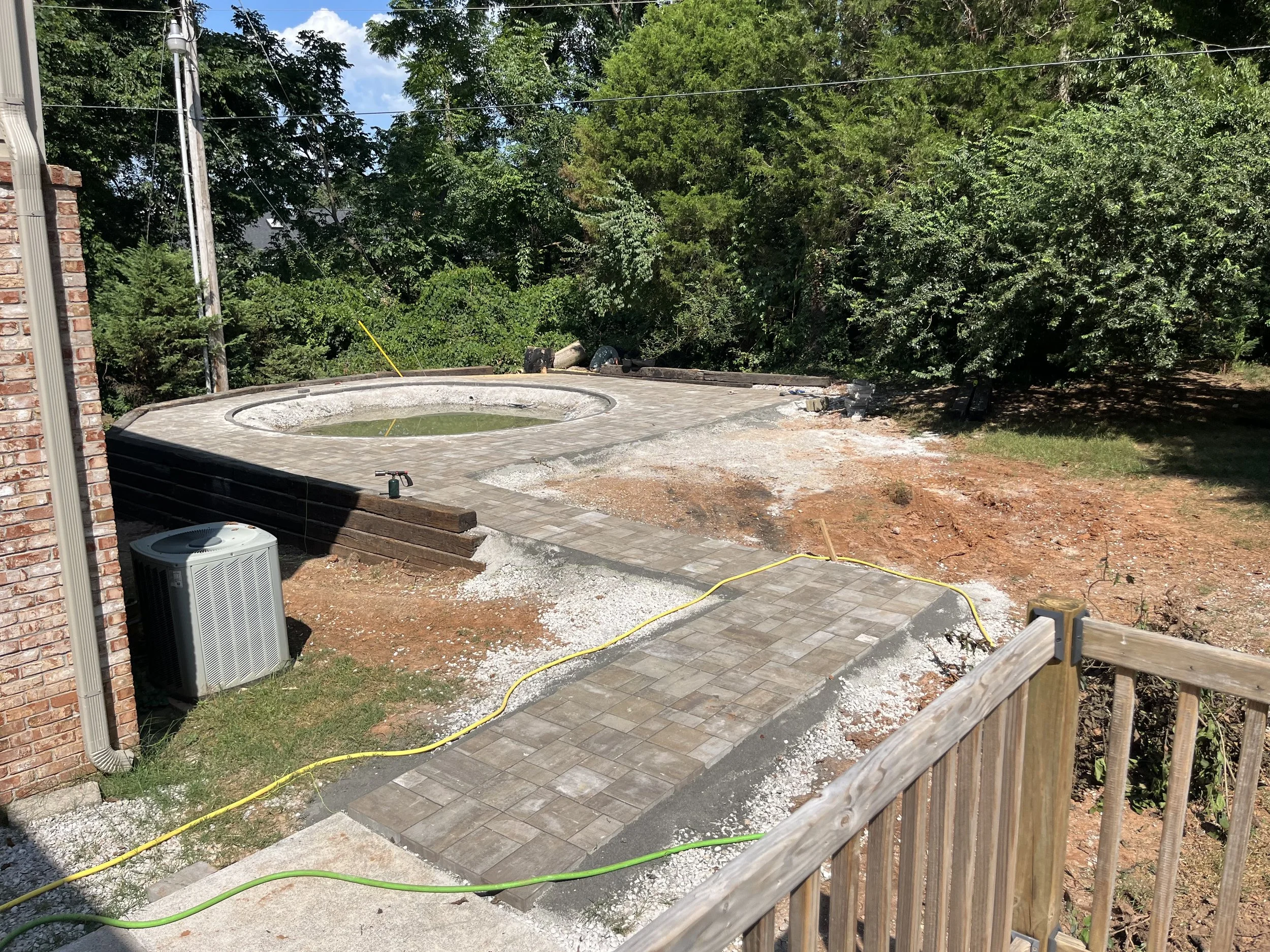Backyard with a circular hot tub on a paved stone patio, next to a brick house and surrounded by trees, with construction materials and tools nearby.