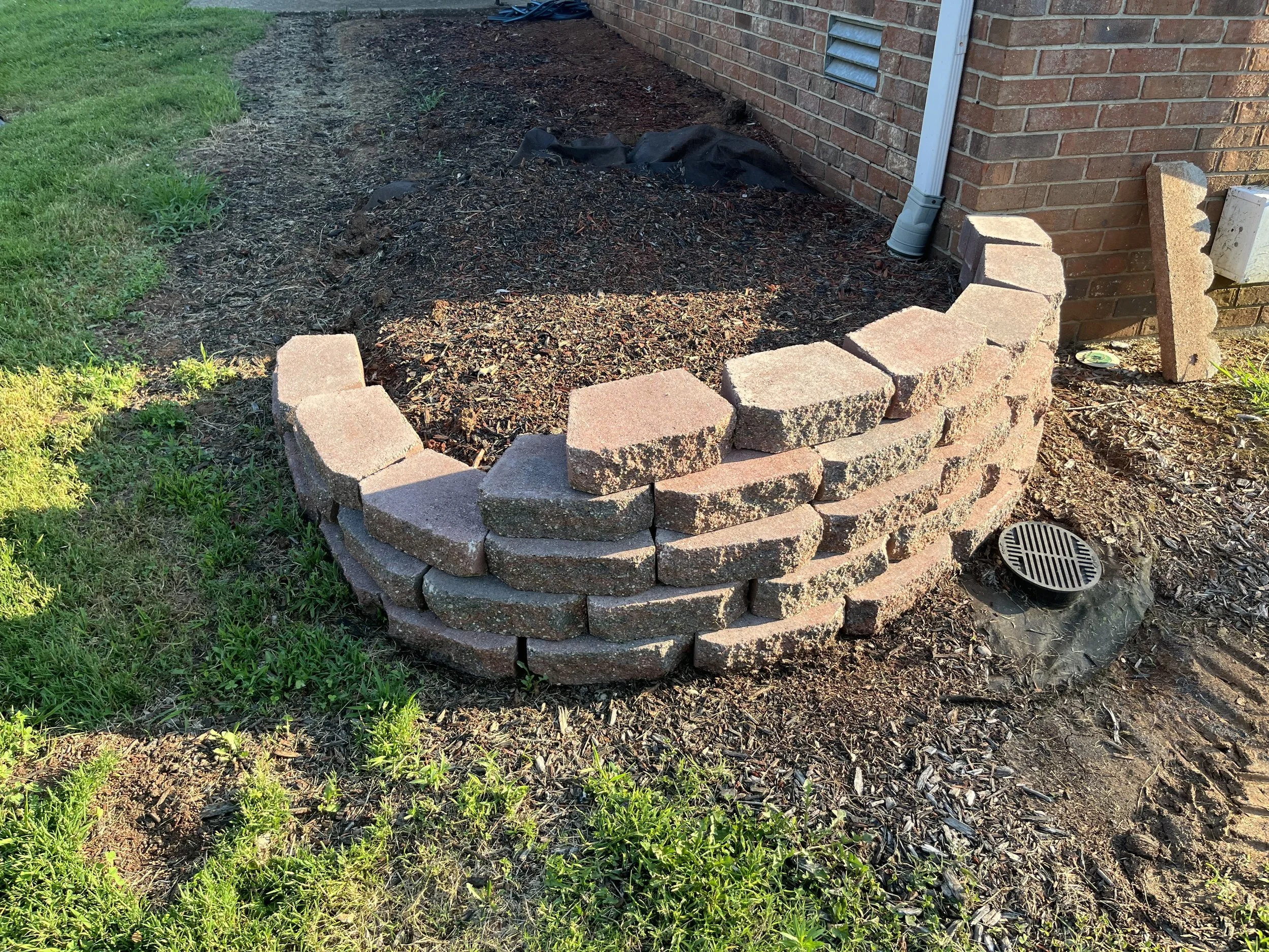 A half-circle brick garden bed with a brick wall at the back, surrounding a small patch of soil, situated next to a brick building with a white downspout. There is a round drain on the ground, and some loose bricks leaning against the building.