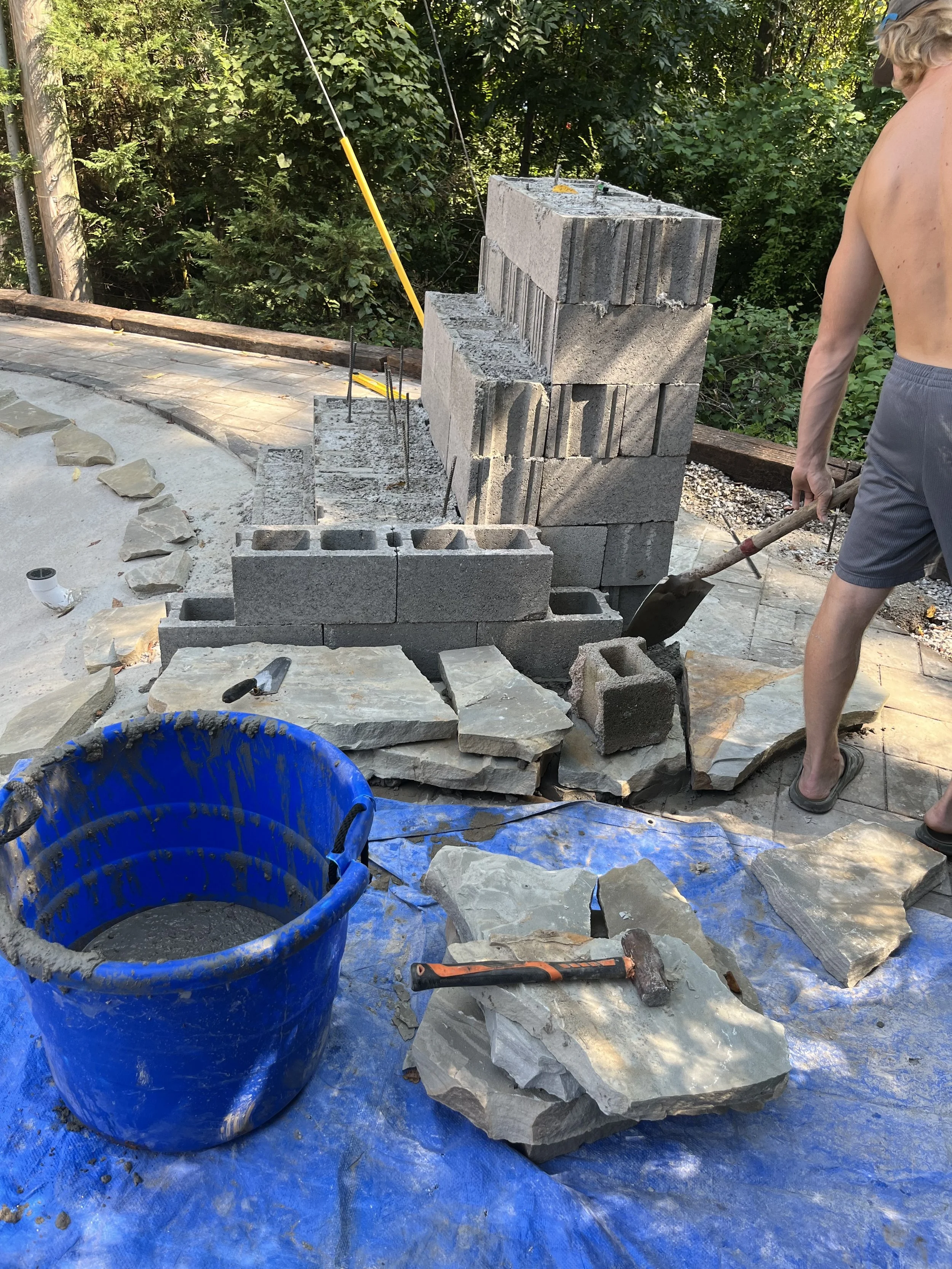 A worker building a stone wall outdoors, with a blue tarp and construction tools around, including a hammer and a bucket of mortar.