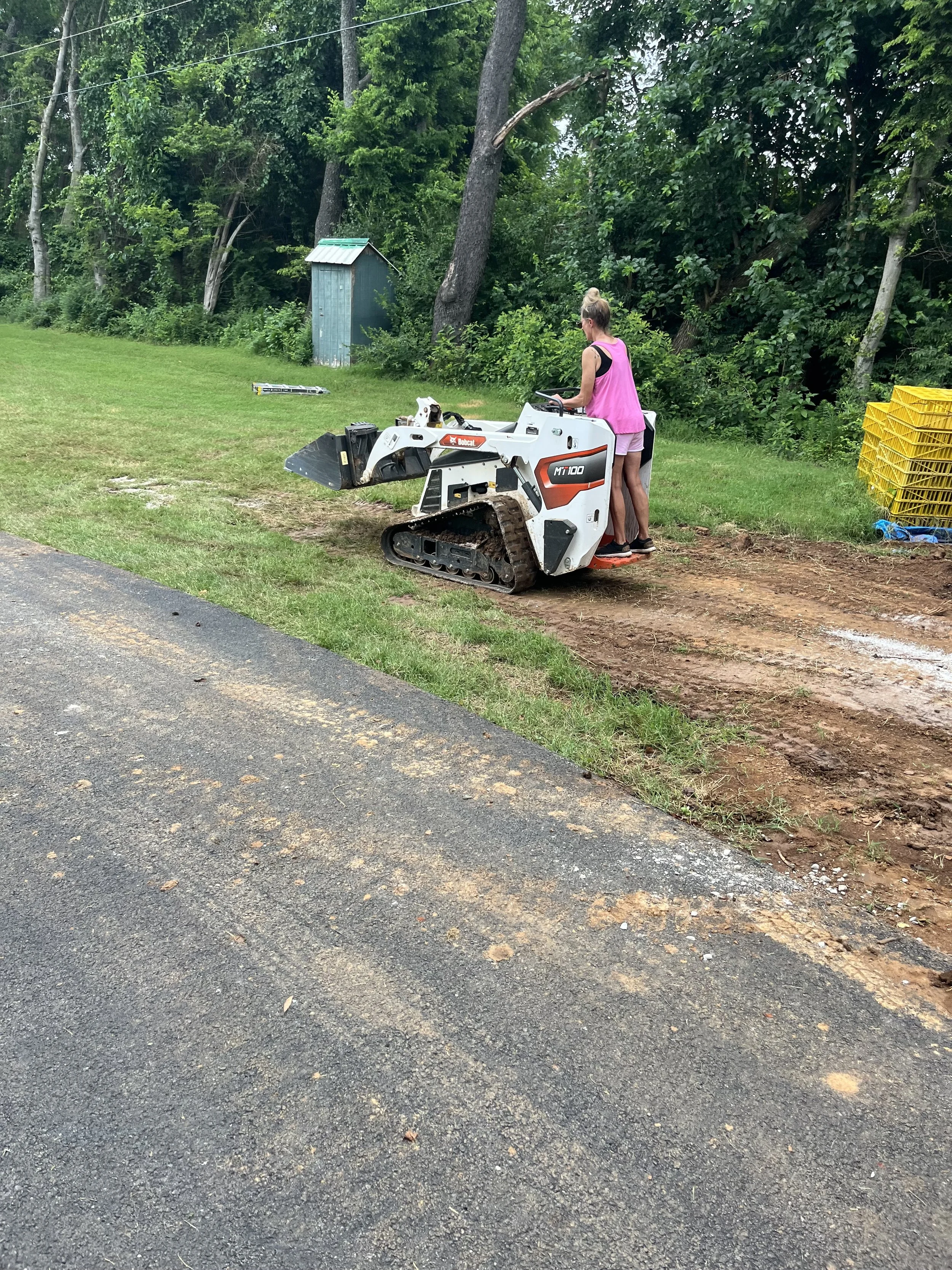 Two women operating a small tracked construction vehicle on a dirt patch next to a grassy area with trees, a small shed, and yellow crates stacked in the background.