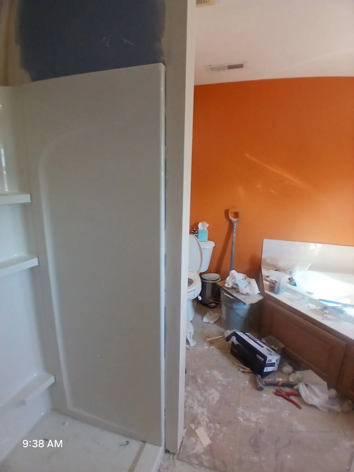 Bathroom with an orange wall and a white toilet, with the shower door open showing a frosted glass shower. The floor and surfaces are messy with tools, cleaning supplies, and debris.