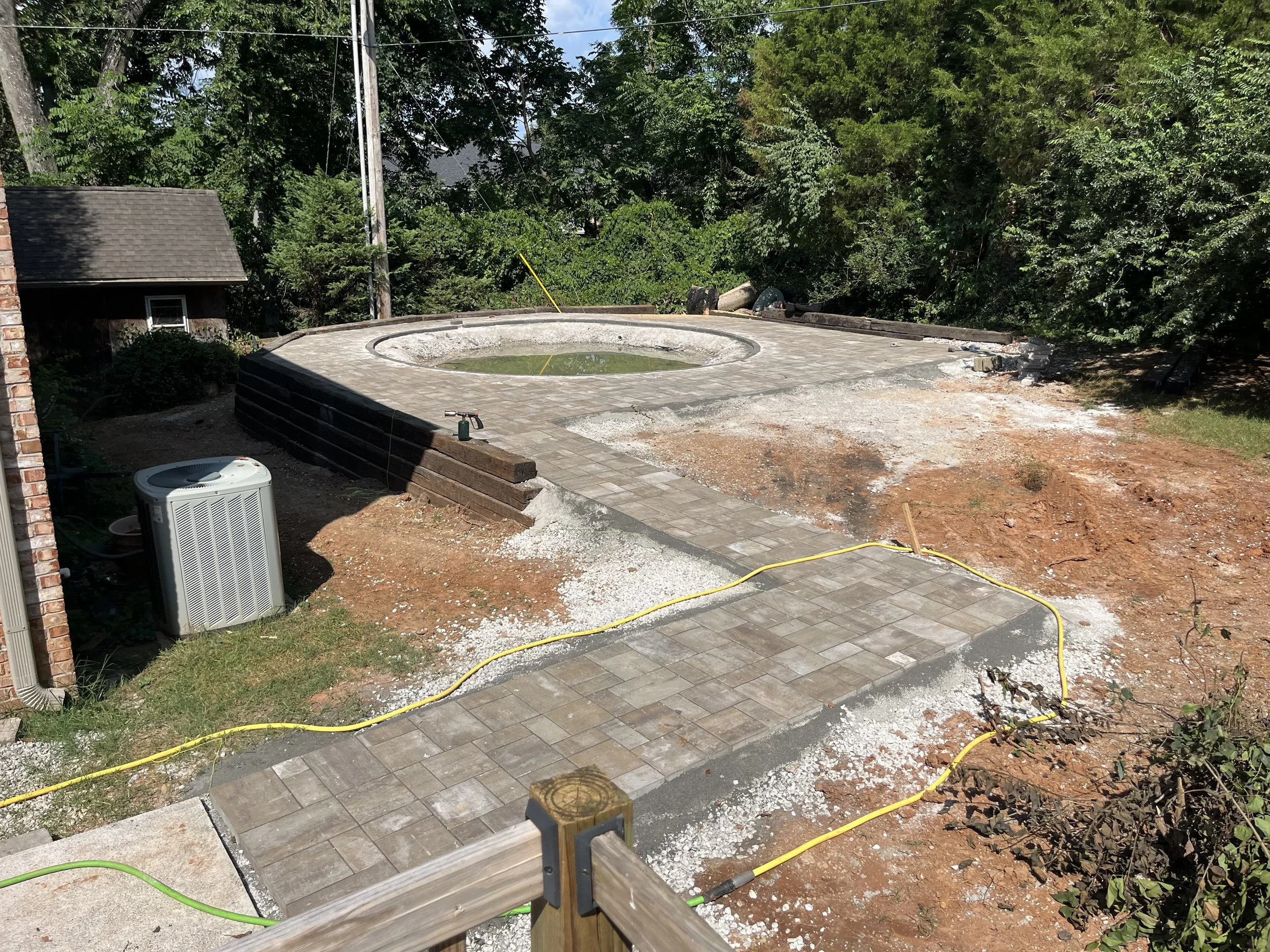 Construction site of a circular swimming pool with brick paving around it. Building materials and tools are visible, with a partially paved pathway. Surrounding greenery and trees in the background.