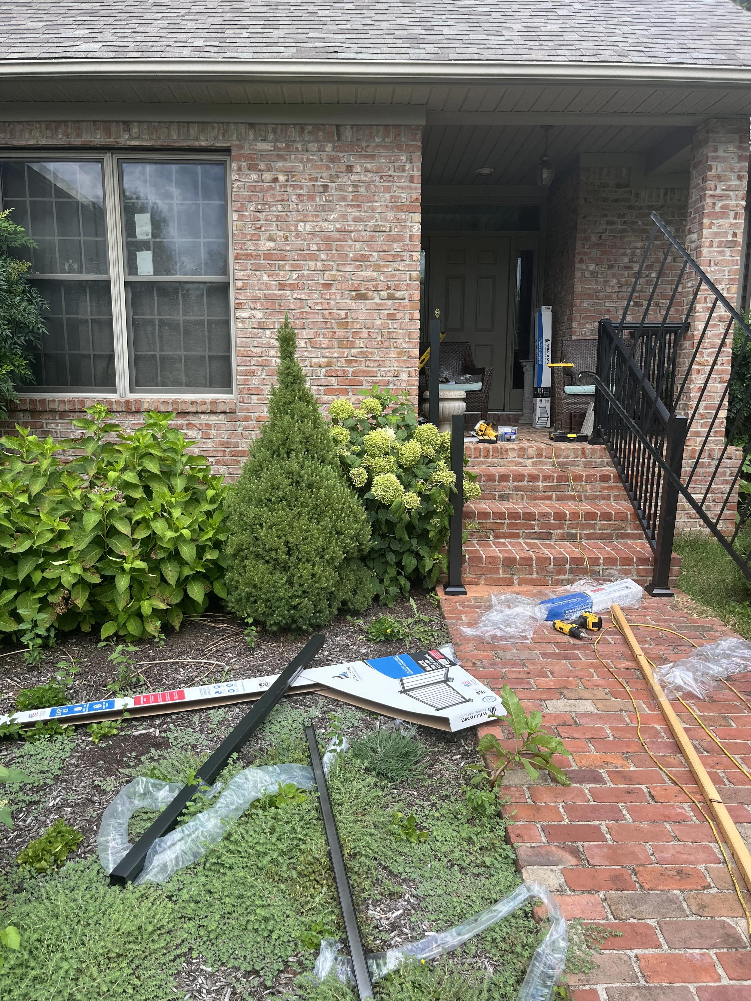 Front porch and steps of a brick house under renovation with construction tools and materials scattered on the brick pathway, and a newly installed black metal railing.