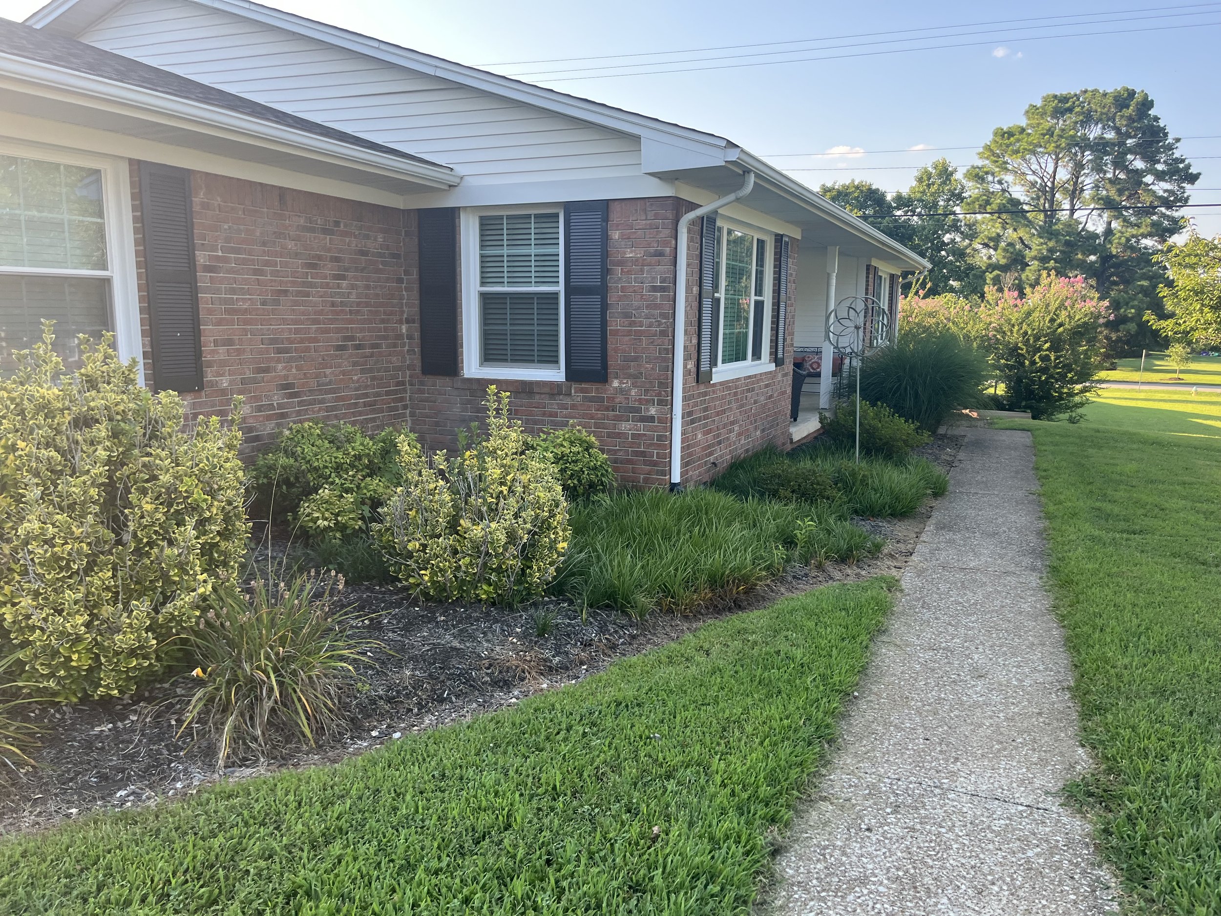 A brick house with white siding, black shutters, a pathway in front, and green landscaping with bushes and trees.