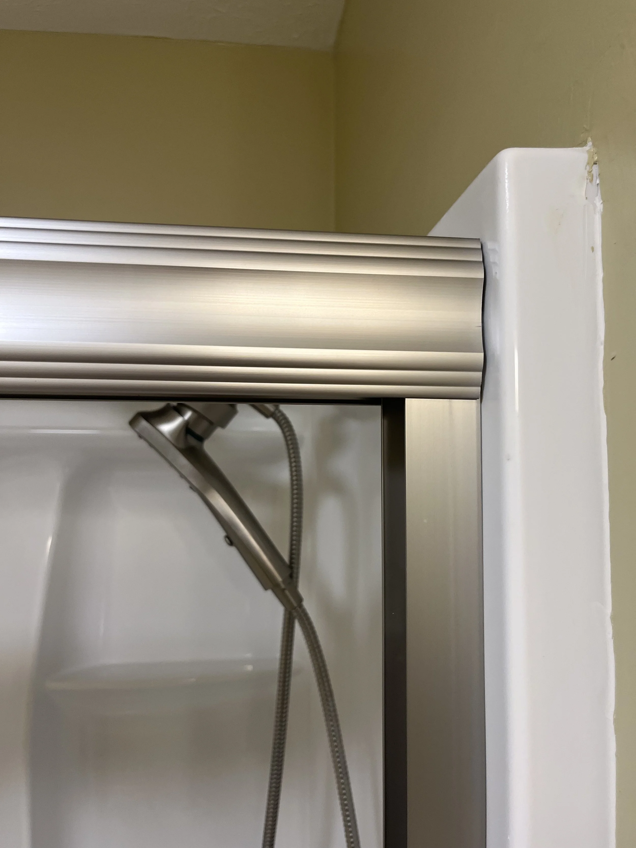 Close-up of a shower stall corner with a silver shower head, flexible hose, and metal frame, with a white shower surround and green wall background.