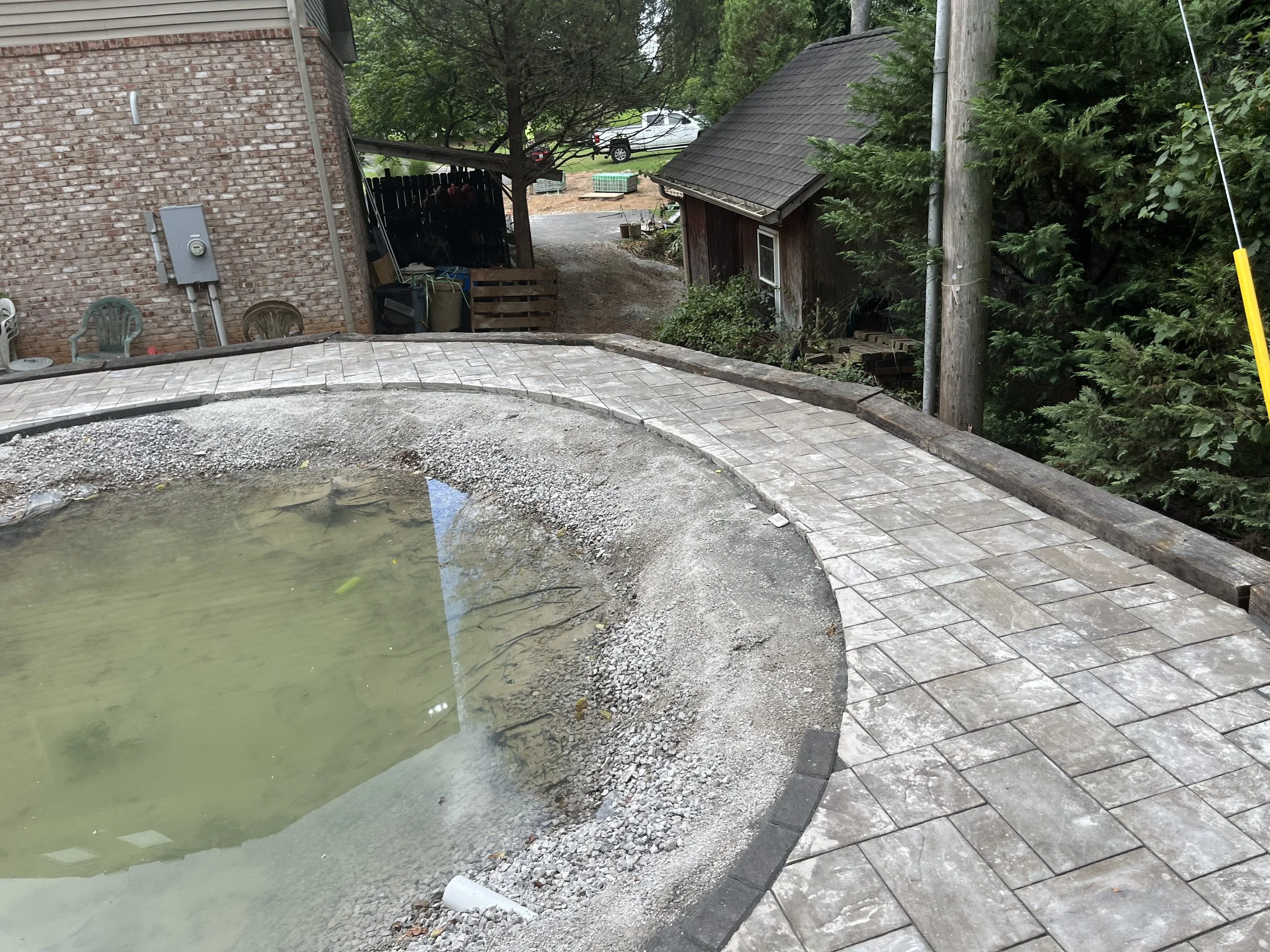 A partially completed stone walkway around an empty pool in a backyard.
