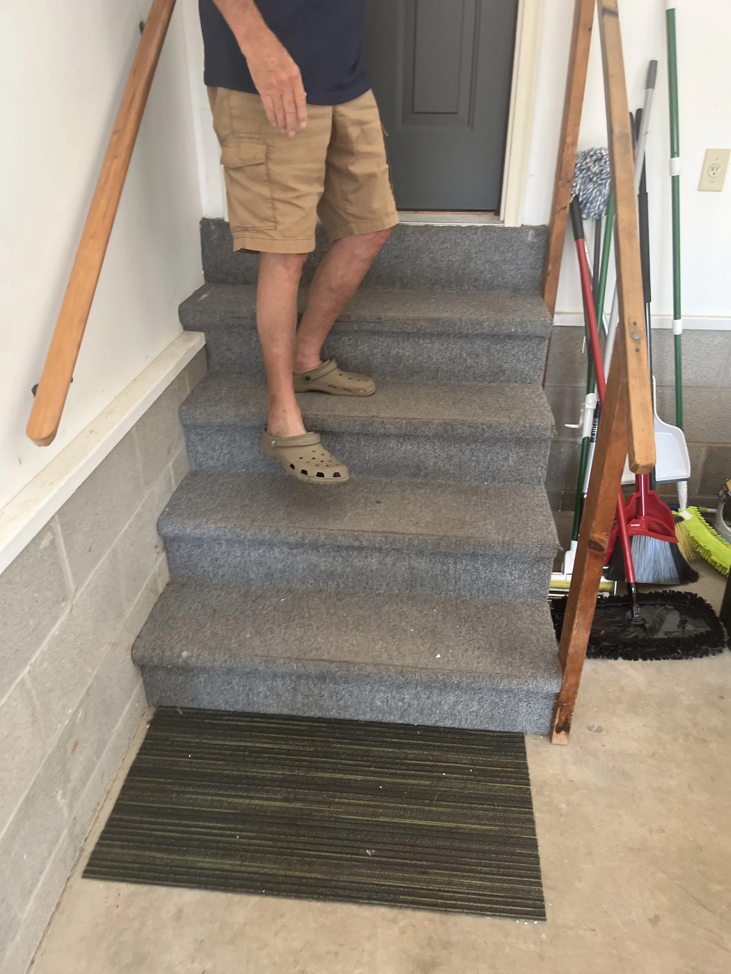Person standing on indoor staircase covered with gray carpet, near cleaning tools and a black door.