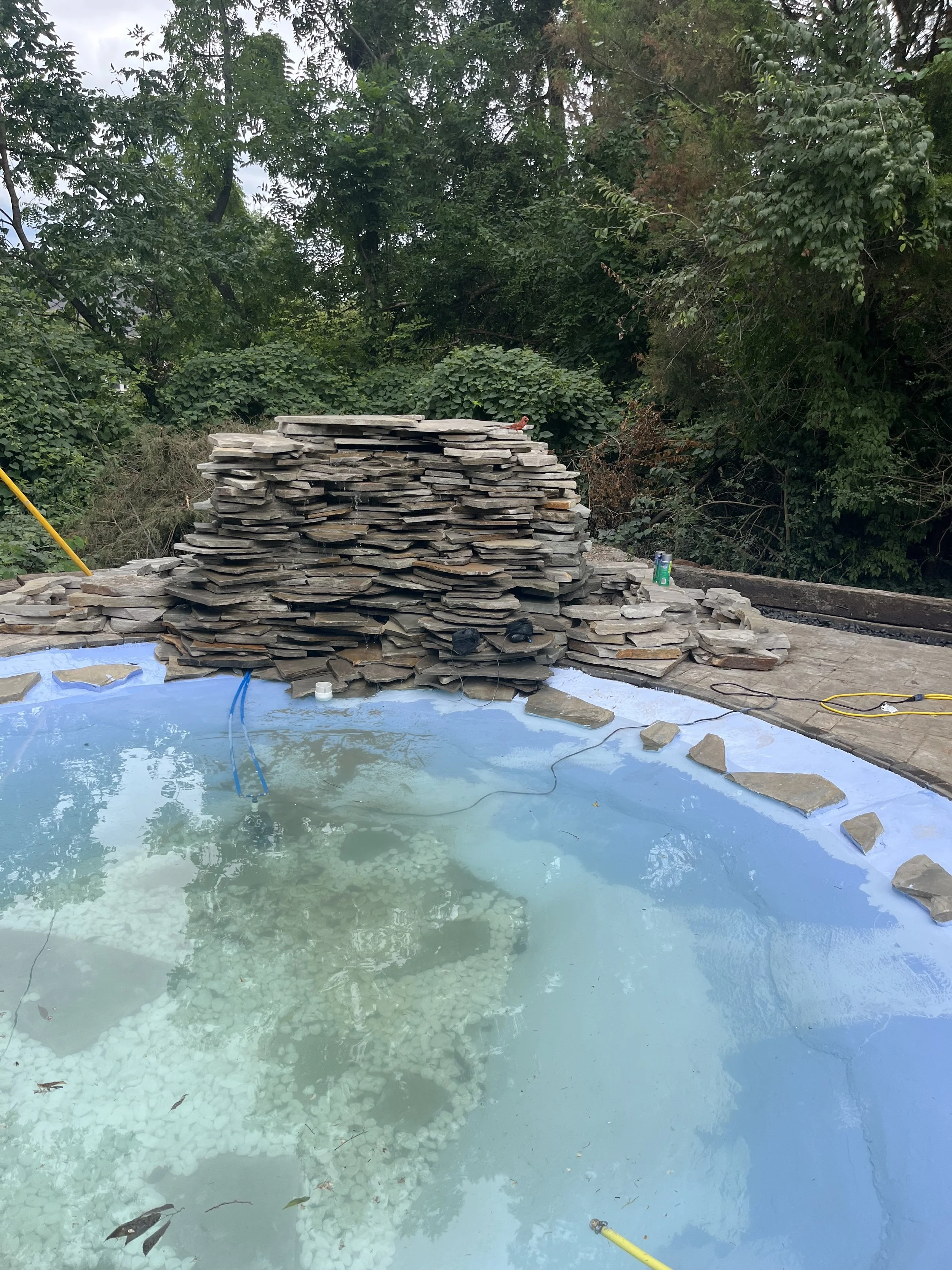 A swimming pool under construction or renovation with a large stack of flat stones piled beside it, surrounded by trees and greenery.