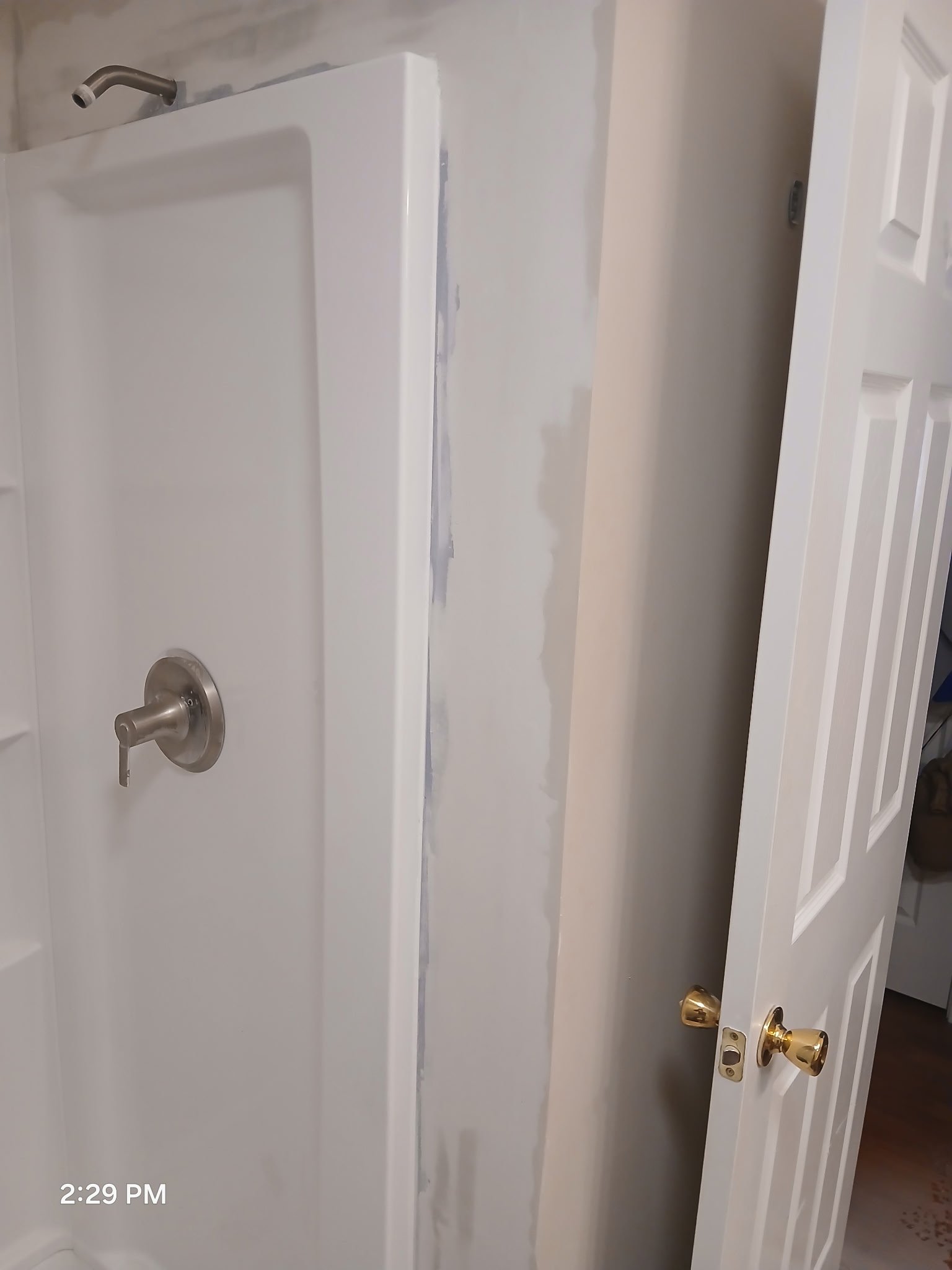 A shower stall with a metal handle and faucet, next to a partially open closet door with gold knobs, and an unfinished wall with visible drywall seam.