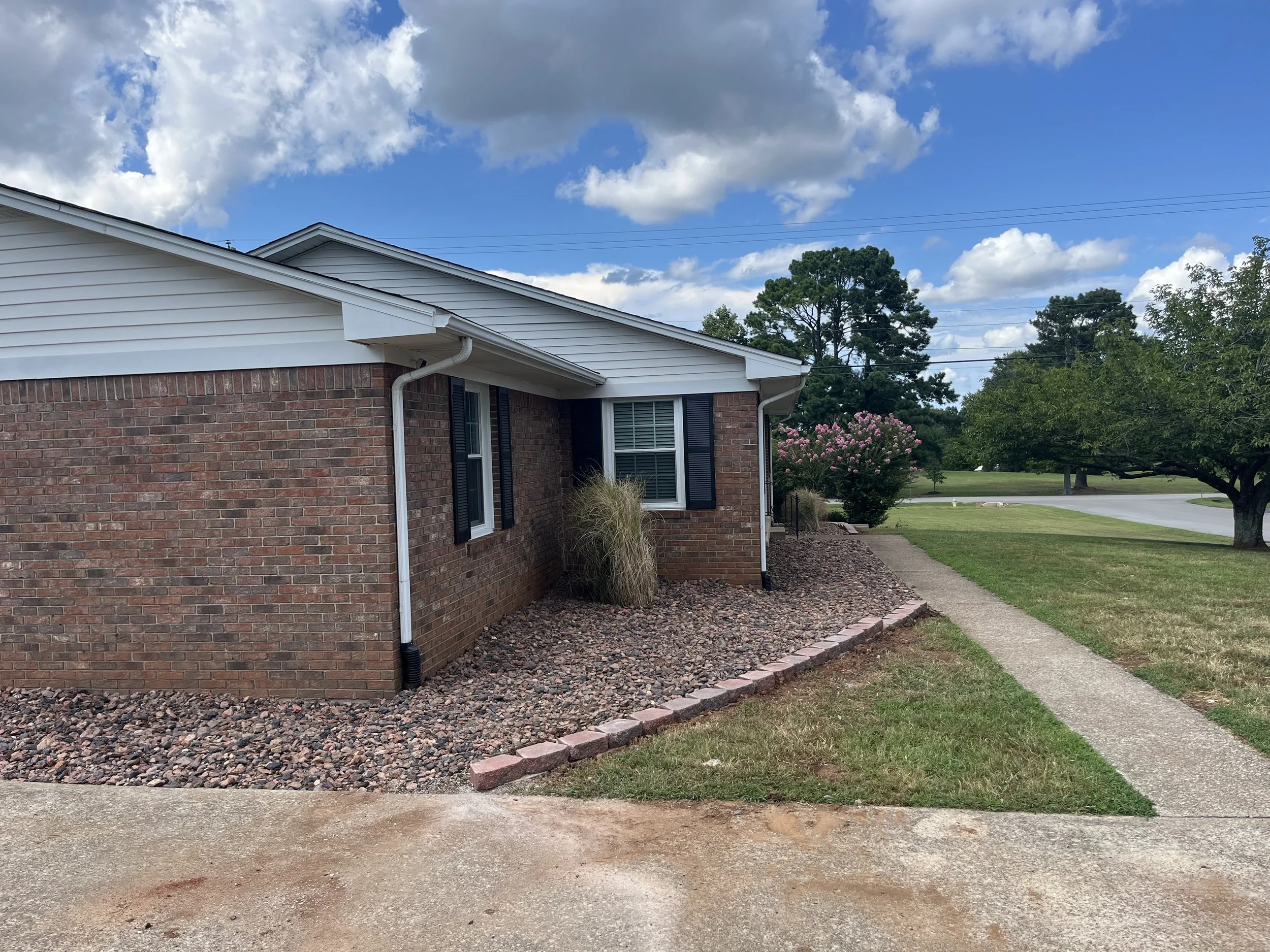 Side view of a brick house with white siding on the roof, black shutters on the windows, and a small front yard with a sidewalk, grass, trees, and a pink flowering bush under a partly cloudy sky.