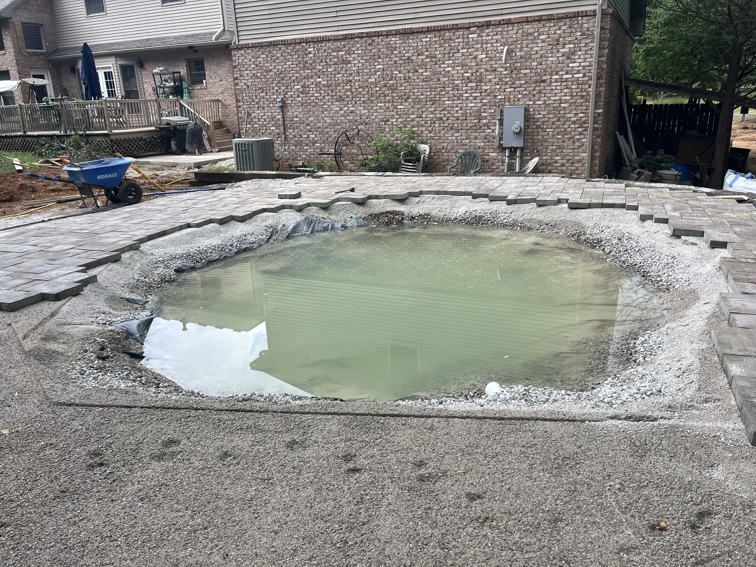 Construction site with removed pool, exposing water and unfinished surrounding patio, in backyard of brick house.