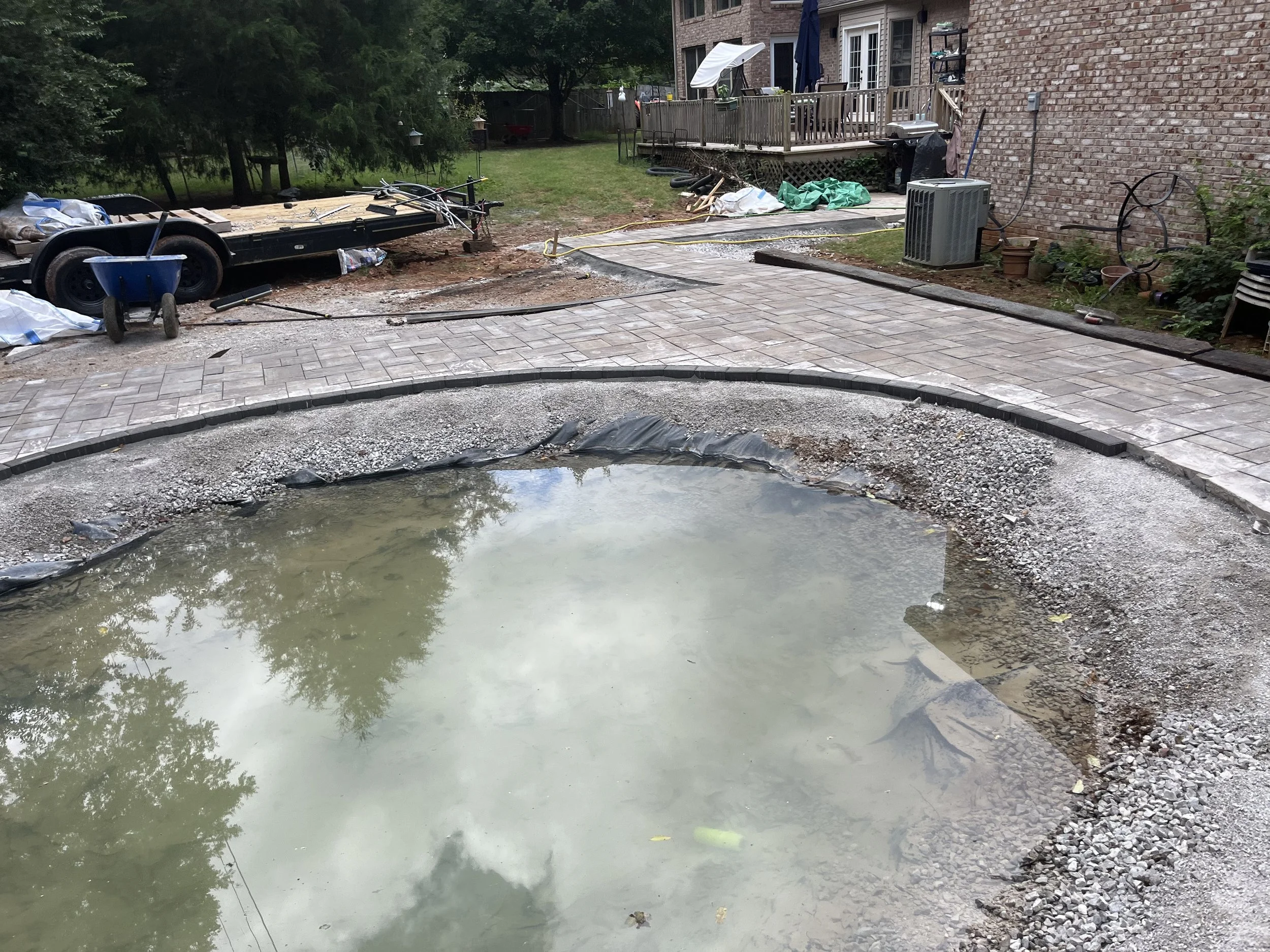 Backyard patio under construction with a partially completed brick paver walkway, a small pond or pool with still water, construction tools and materials, a trailer, and a house with a deck in the background.
