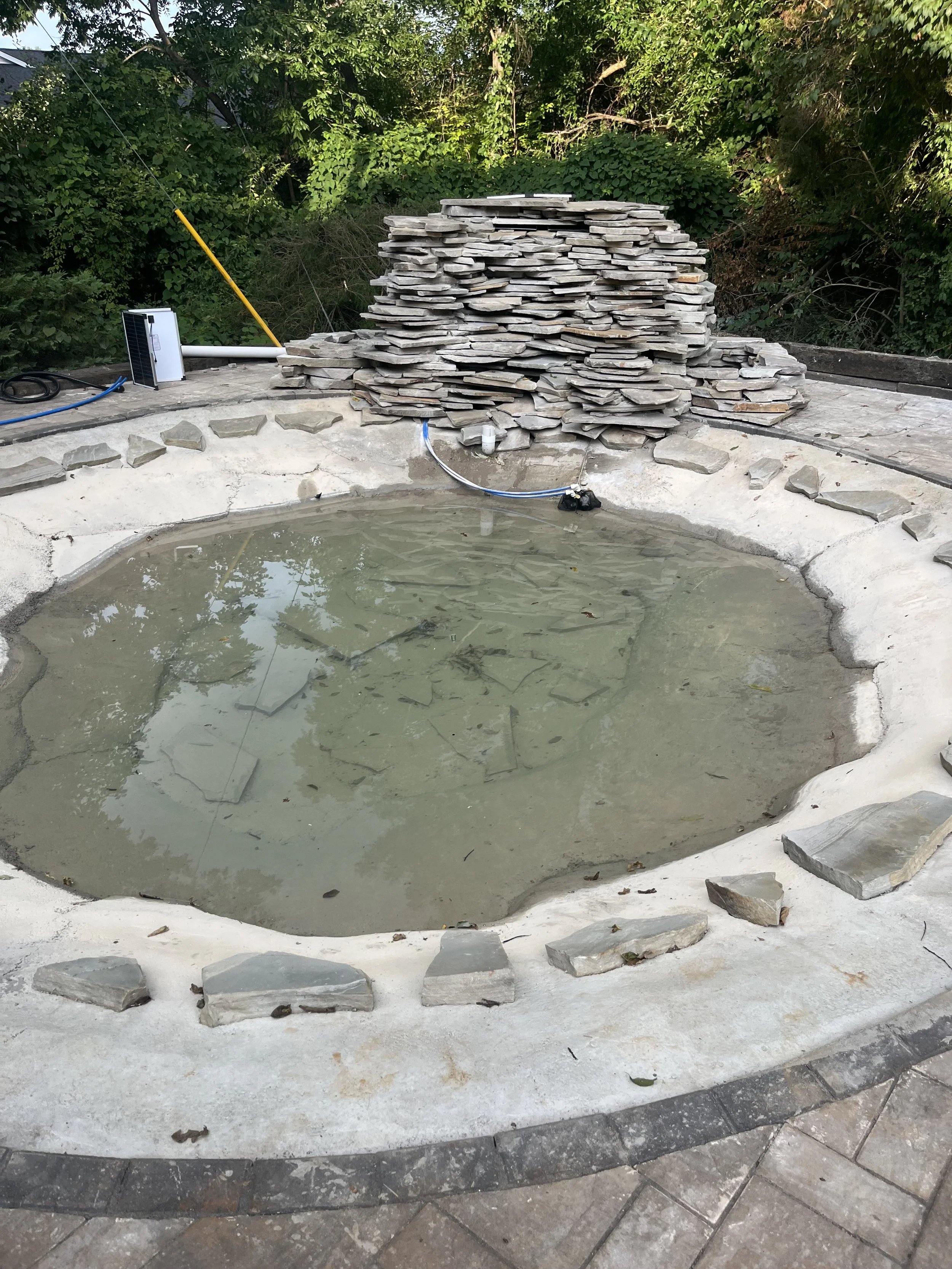 A small, unfinished outdoor pool surrounded by a stone and concrete border, with a tall pile of flat stones and outdoor equipment nearby, set in a backyard with trees and greenery.