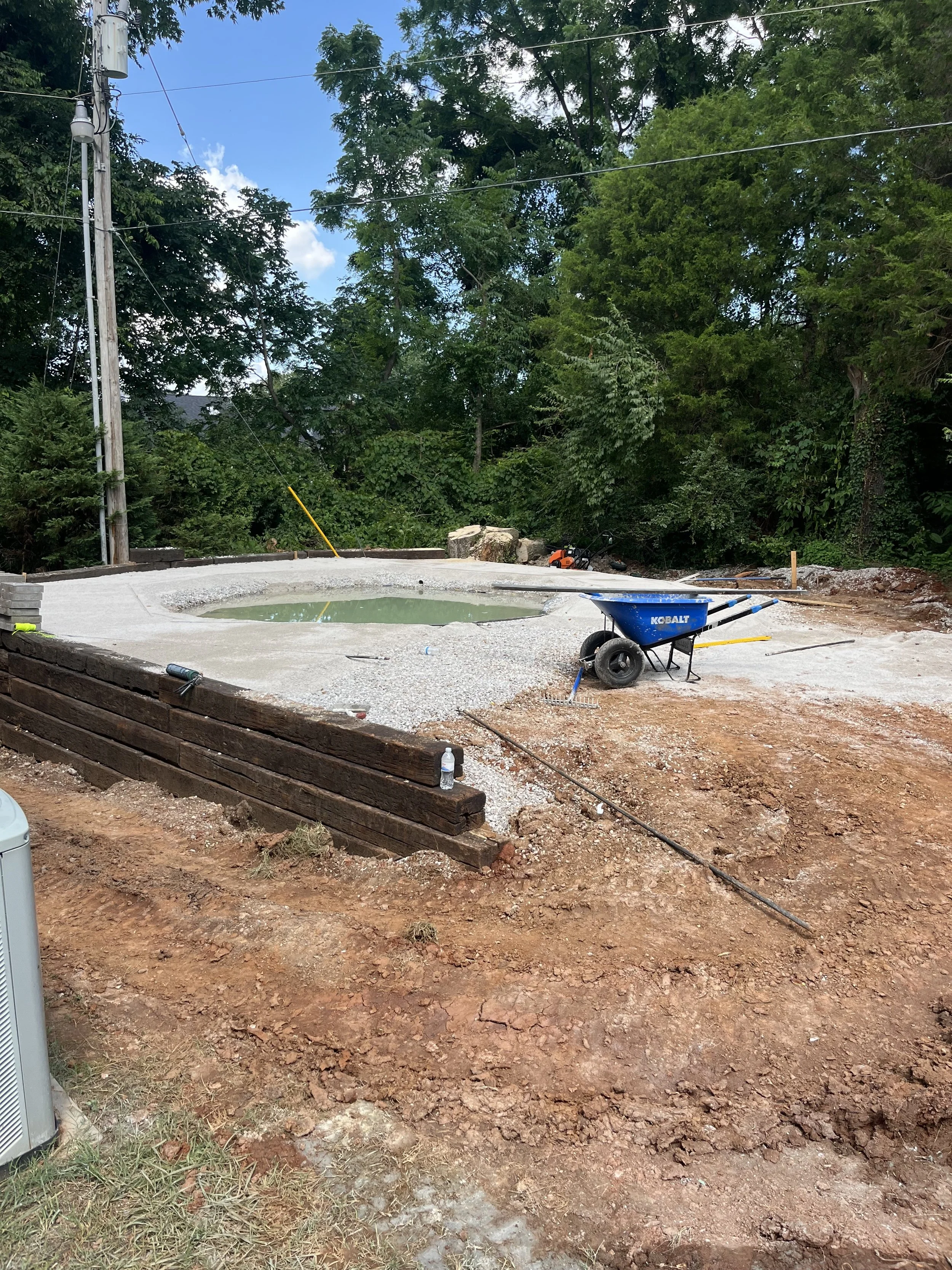 Under construction backyard with a small pond, a blue wheelbarrow, and wooden border in an outdoor setting surrounded by trees.