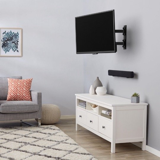 Living room with a gray couch, patterned throw pillow, beige ottoman, white media console with vases and decor, wall-mounted TV, soundbar, framed artwork, and a rug on light wood flooring.