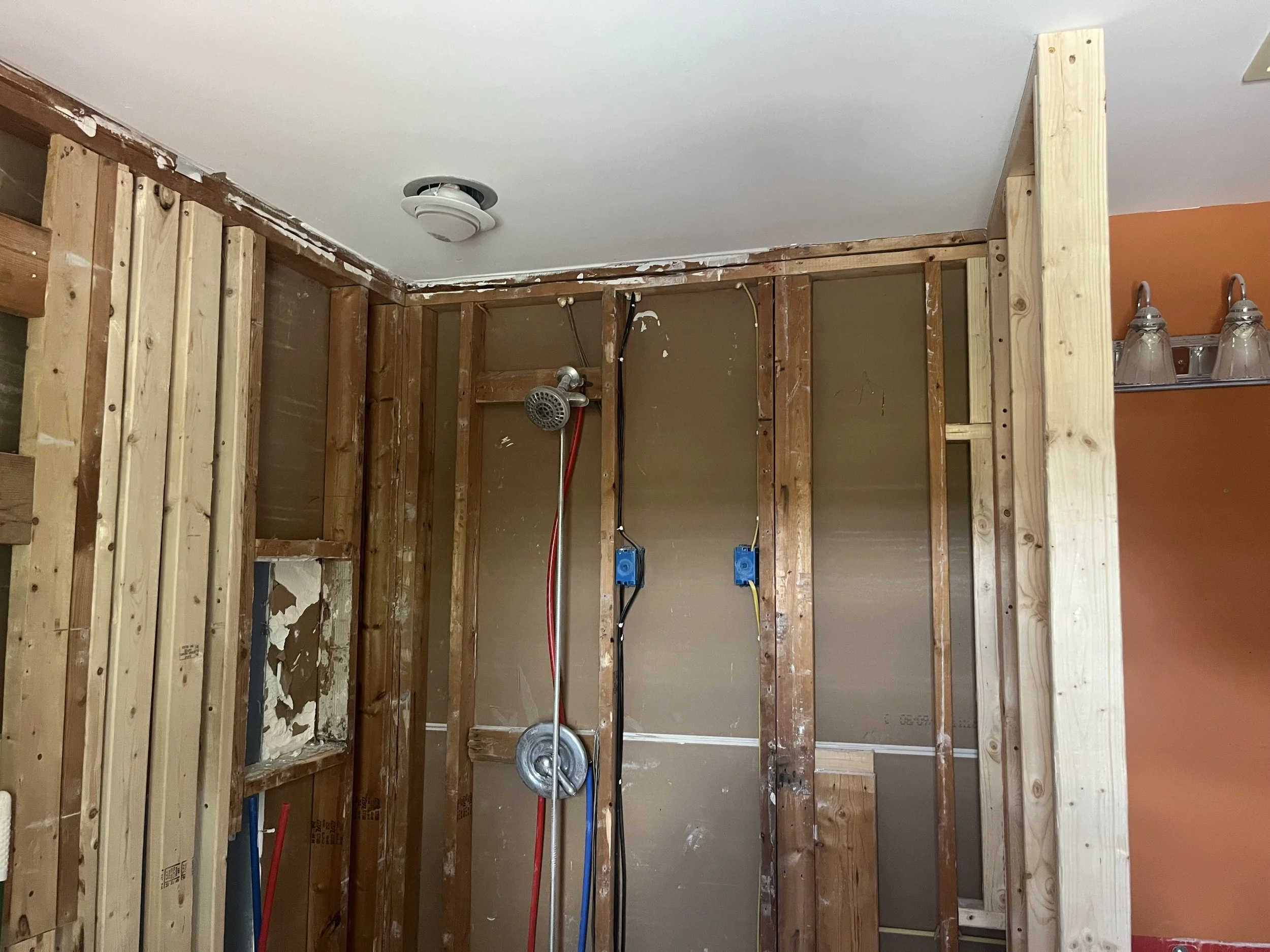 A bathroom under renovation with exposed wooden studs, a ceiling smoke detector, and plumbing pipes.