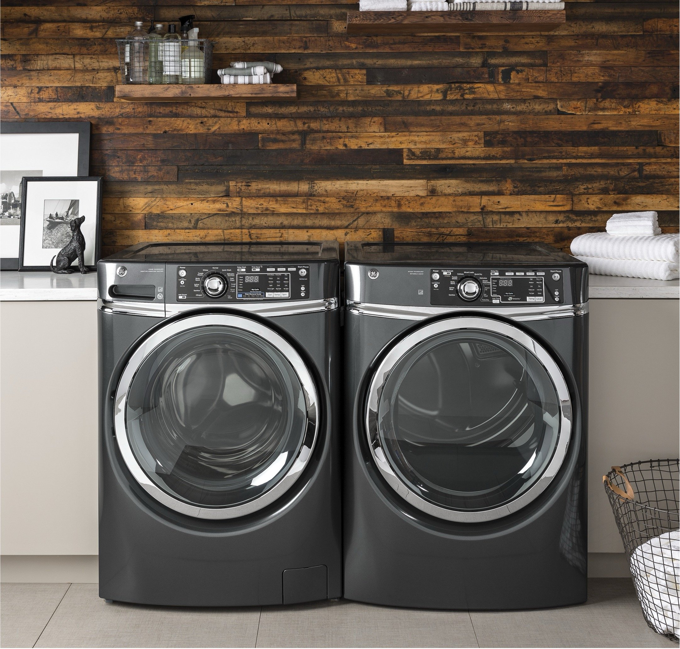 A modern laundry room with a dark gray front-loading washer and dryer set, set against a wooden plank wall. The countertop is decorated with framed photographs, a small black dog figurine, and neatly folded white towels, with a wire laundry basket nearby.