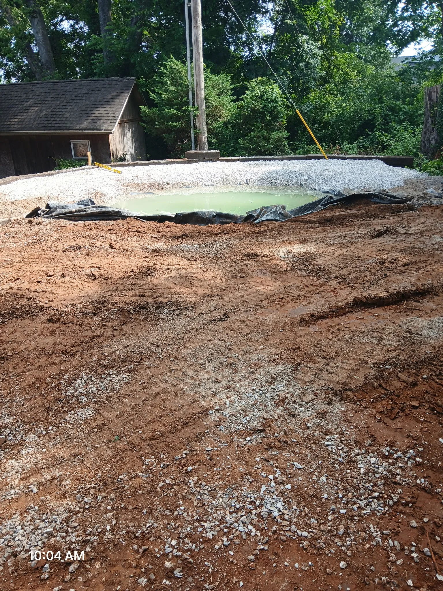 Construction site with a newly built or in-progress small pond or pool, surrounded by red dirt and gravel, with a black tarp lining part of it, and a wood shed and trees in the background.