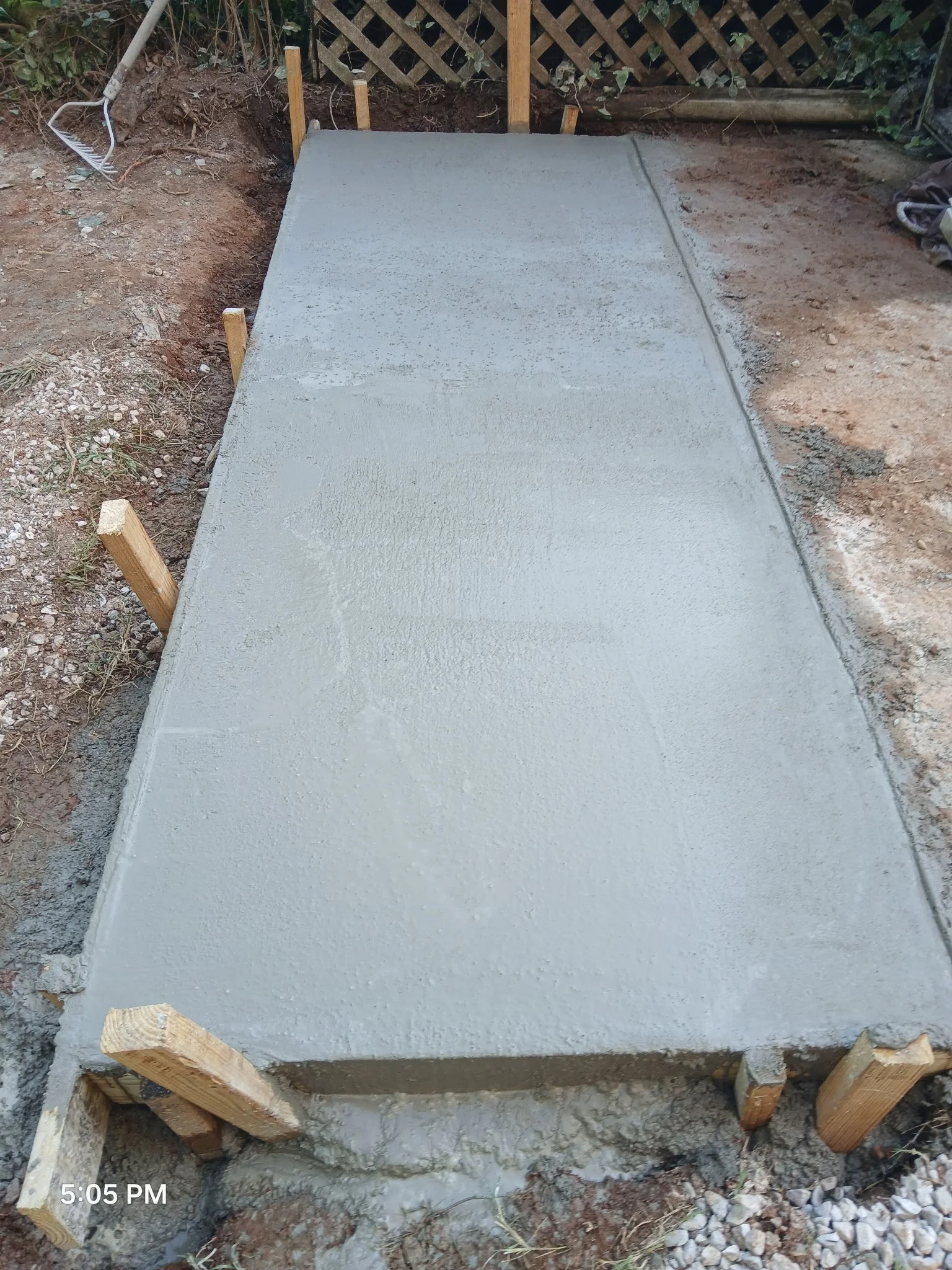 Freshly poured concrete slab on a construction site, bordered by wooden stakes with a gravel patch at the bottom right and gardening tools nearby.
