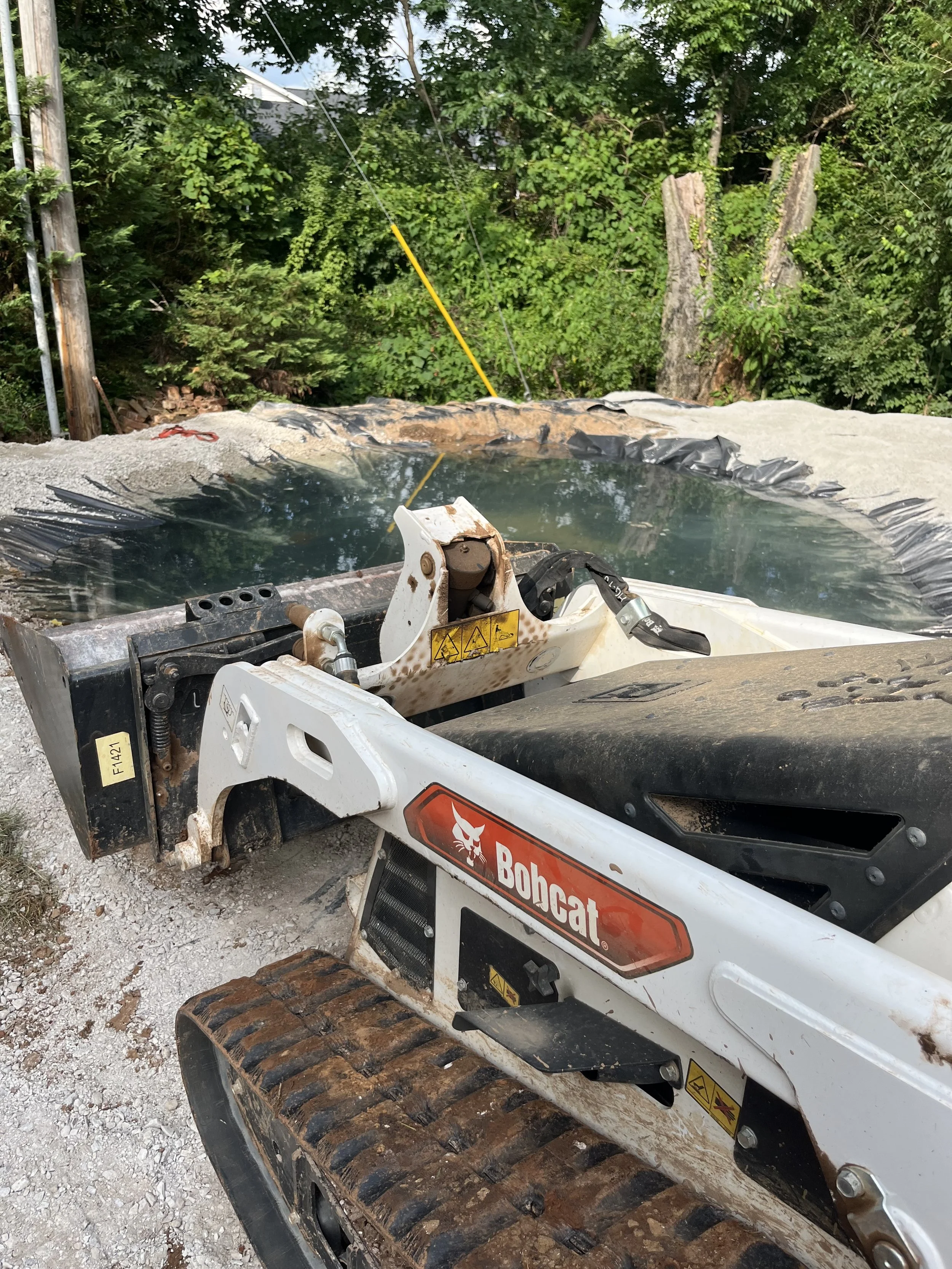A Bobcat mini excavator with a label, working on a construction site with a pond or water feature, surrounded by gravel and greenery.