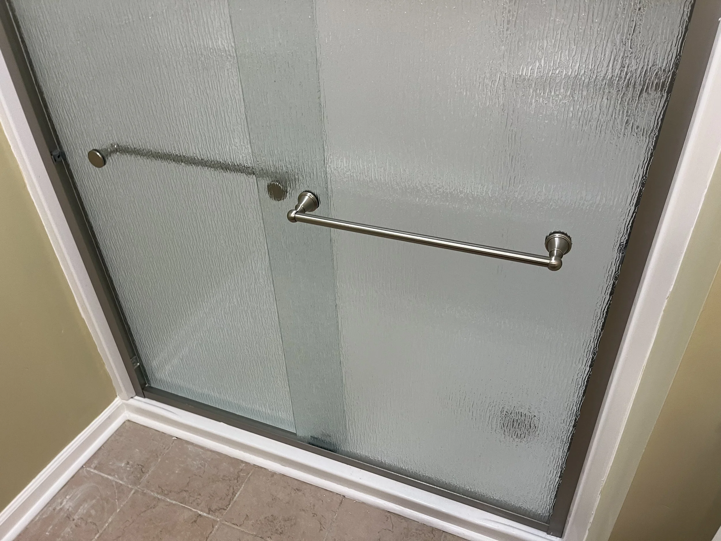 A glass shower door with a silver towel bar installed on it, located in a bathroom with beige walls and tiled flooring.