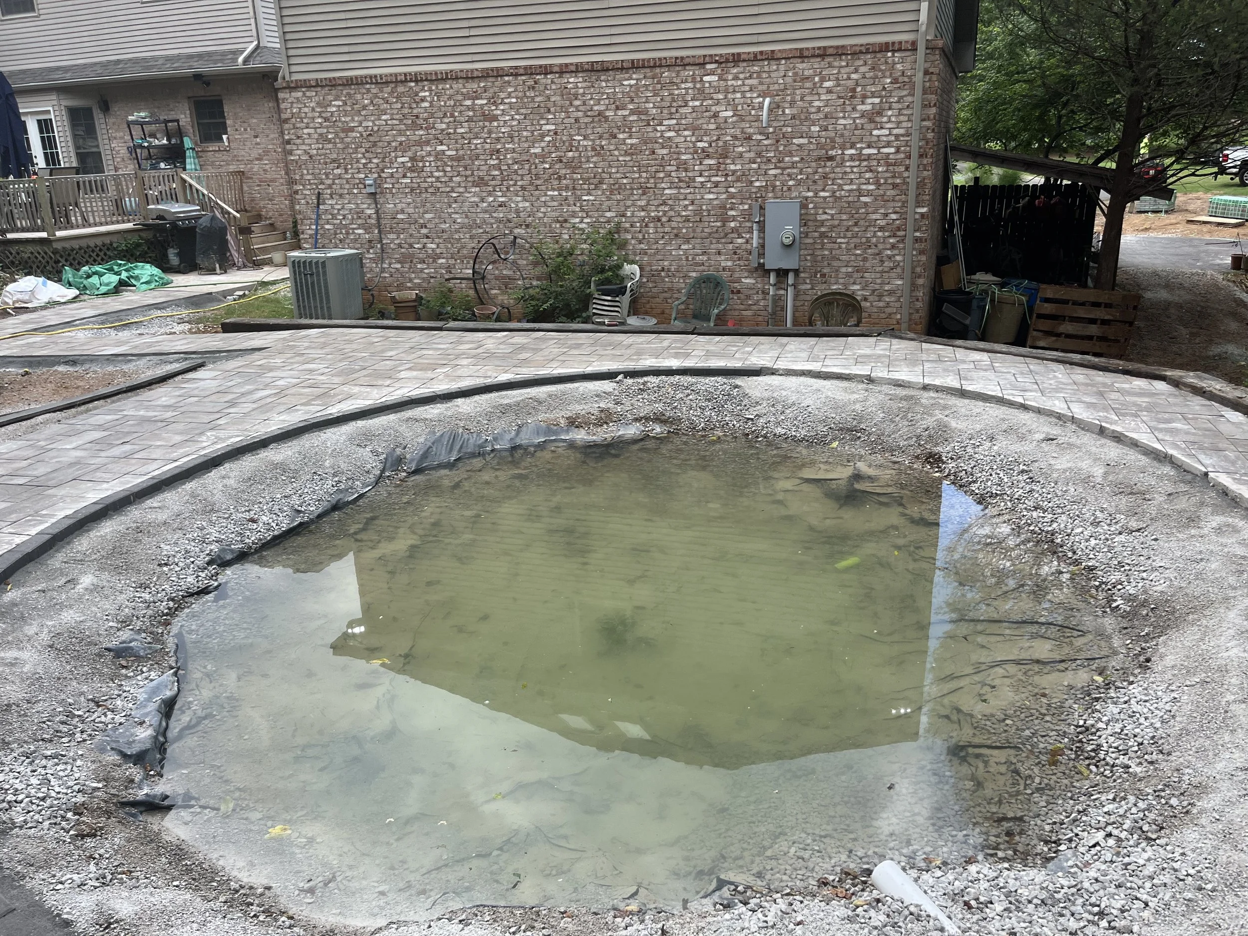 A small, round, unfinished concrete pool filled with water in a backyard, with a partially completed brick pathway surrounding it.