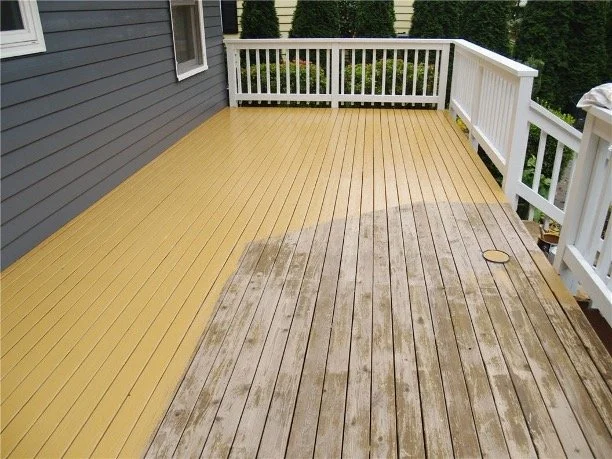 A wooden deck with a section of new yellow stain and an older weathered section next to a house with gray siding and white railings.