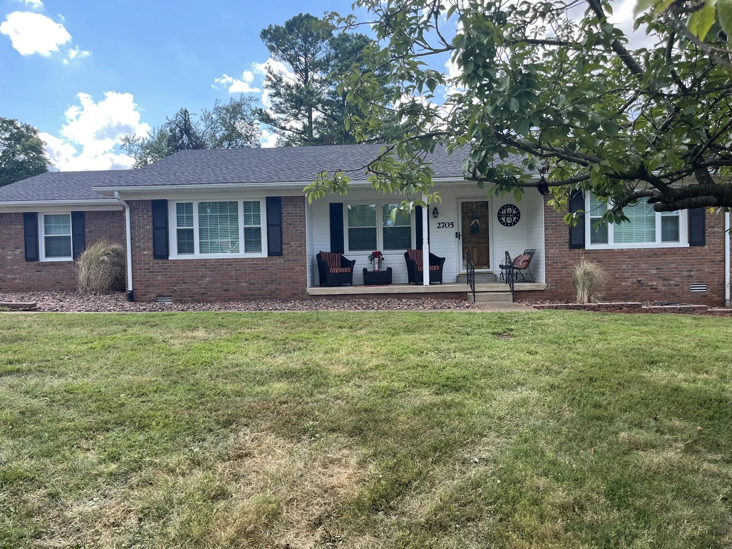 A single-story brick house with a front porch, decorated with outdoor seating, in a yard with a tree and green grass, under a partly cloudy sky.